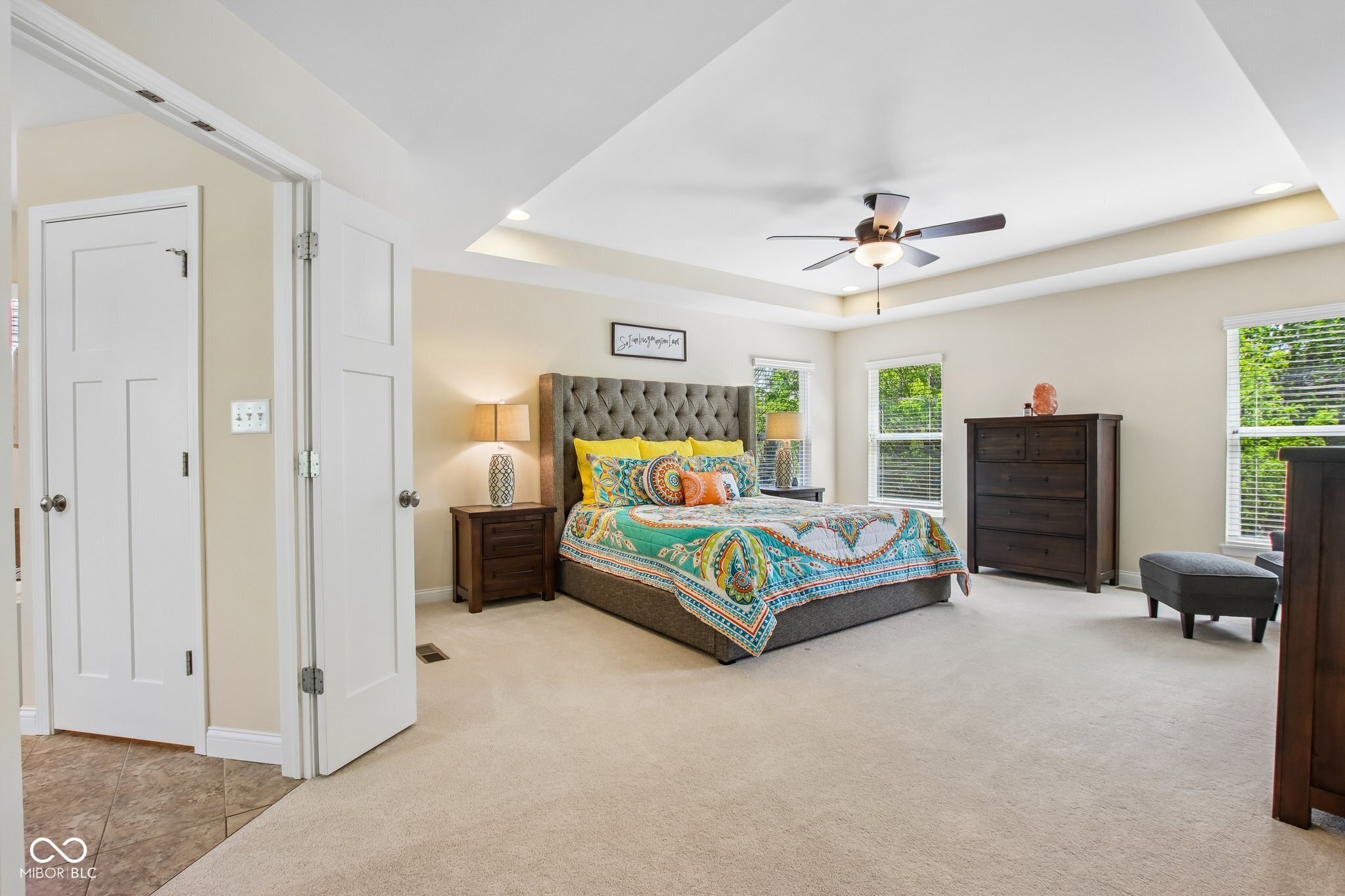 Primary bedroom with colorful patterned bedding, a tufted gray headboard, dark wood dressers, and a tray ceiling.