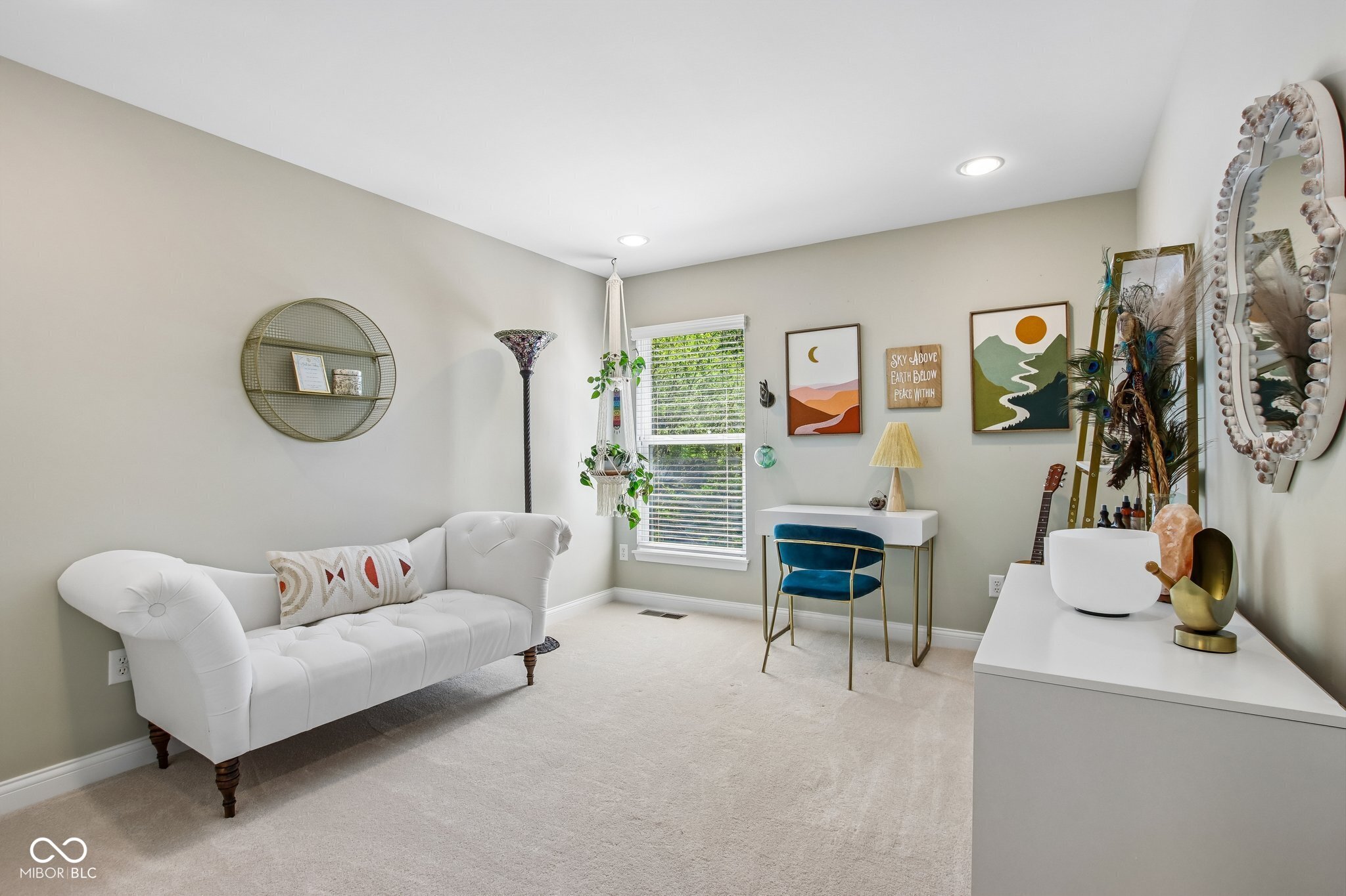 Sitting room with a white tufted loveseat, small desk, blue chair, and colorful landscape artwork.