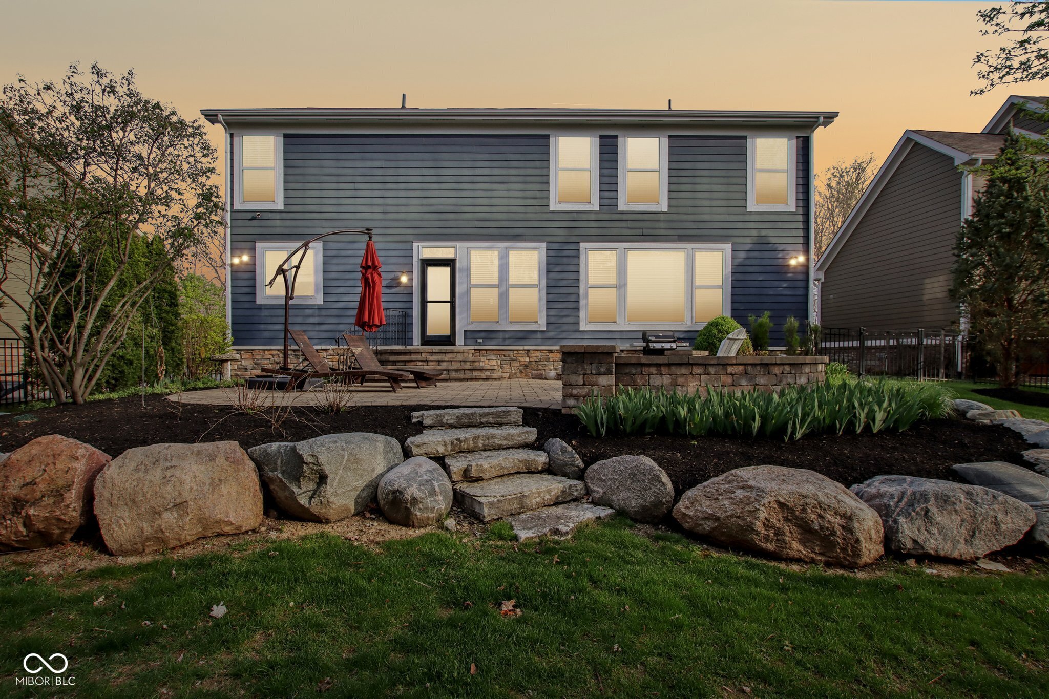 Two-story blue-gray house with a stone patio, outdoor seating, and boulder landscaping at dusk.