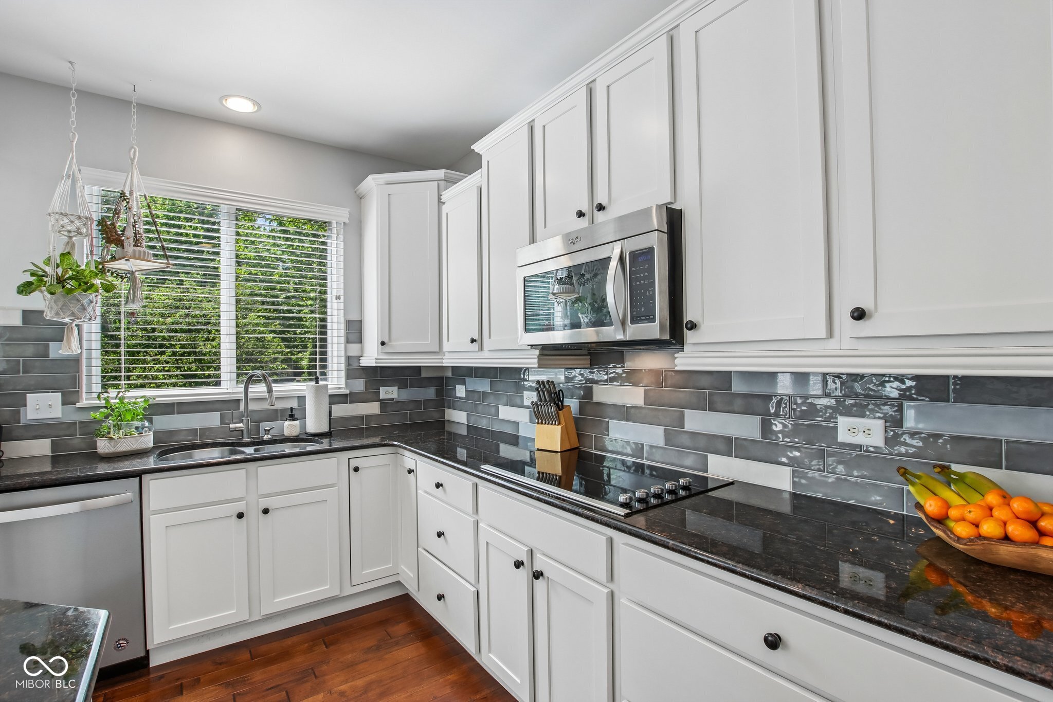 Bright kitchen with white cabinets, dark countertops, gray tile backsplash, and a window overlooking greenery.