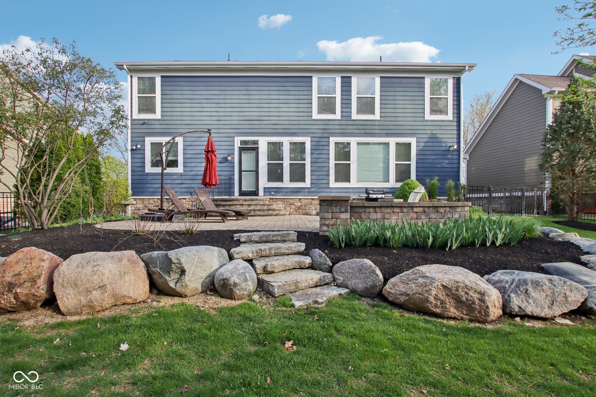 Blue two-story home with a raised stone patio, red umbrella, and landscaped rock garden steps.