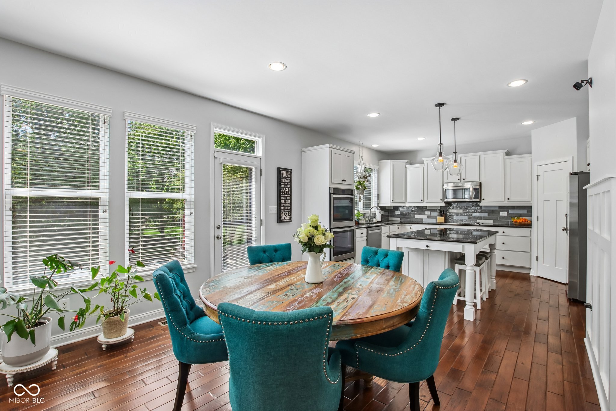 Open kitchen and dining area with white cabinets, teal chairs, and a colorful round wood table.