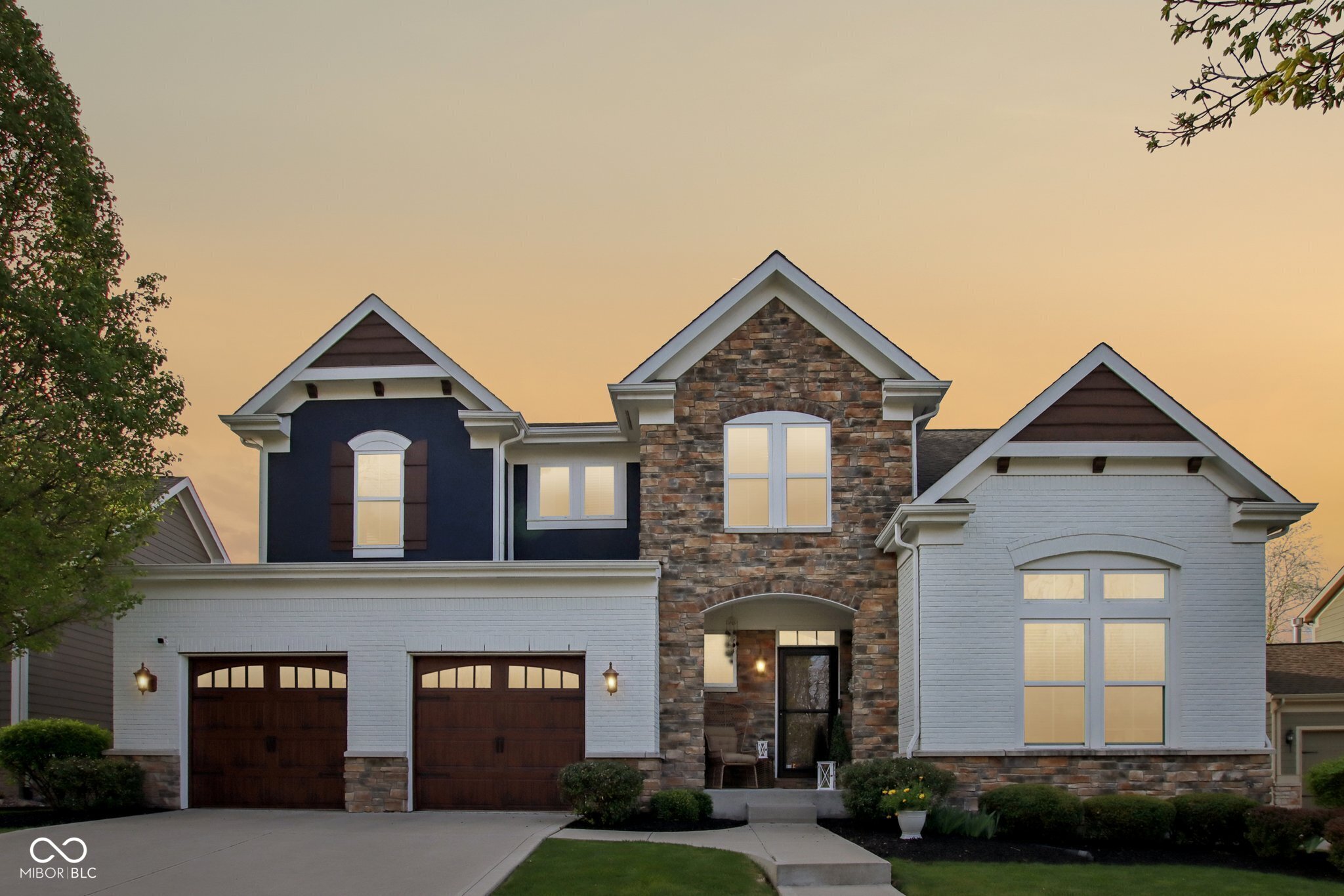 Two-story house with a stone facade, white brick wings, and two dark wood garage doors at sunset.
