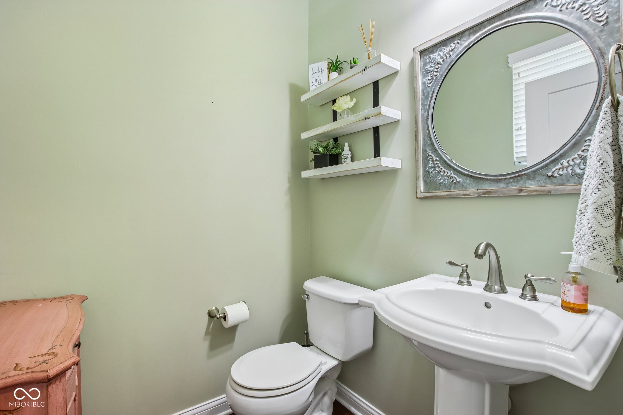 Bathroom with a white pedestal sink, ornate mirror, floating shelves, and pale green walls.
