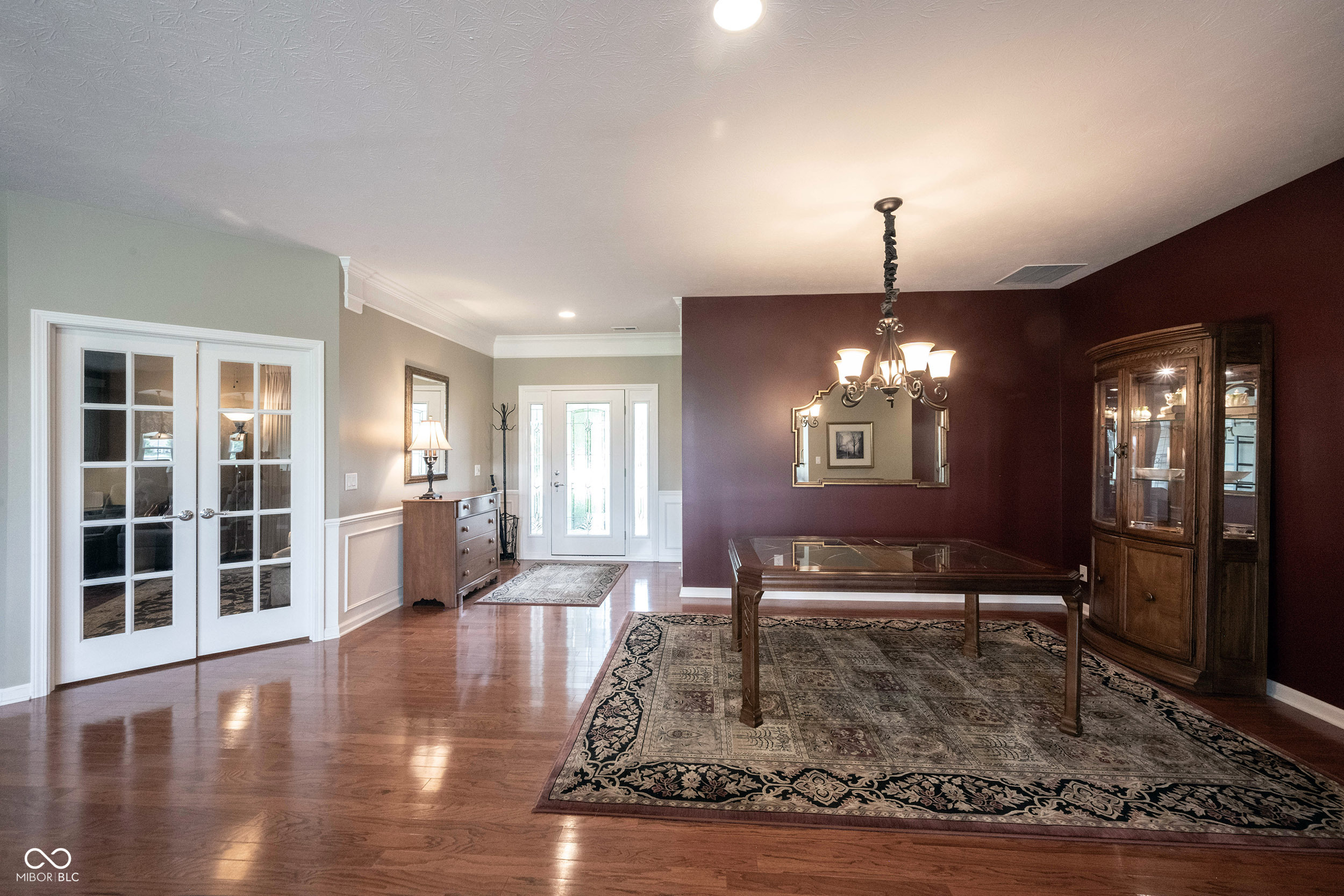 Formal dining room with a glass-top table, chandelier, burgundy walls, and a large patterned rug.