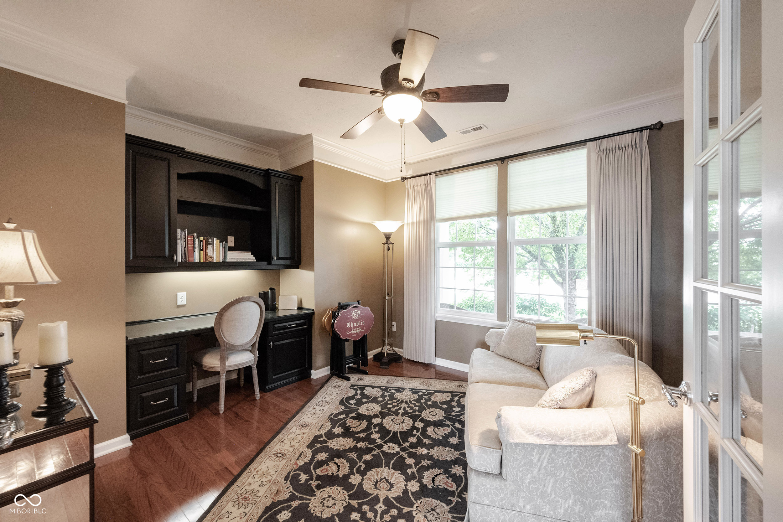 Home office with dark built-in desk, white sofa, and large curtained windows.