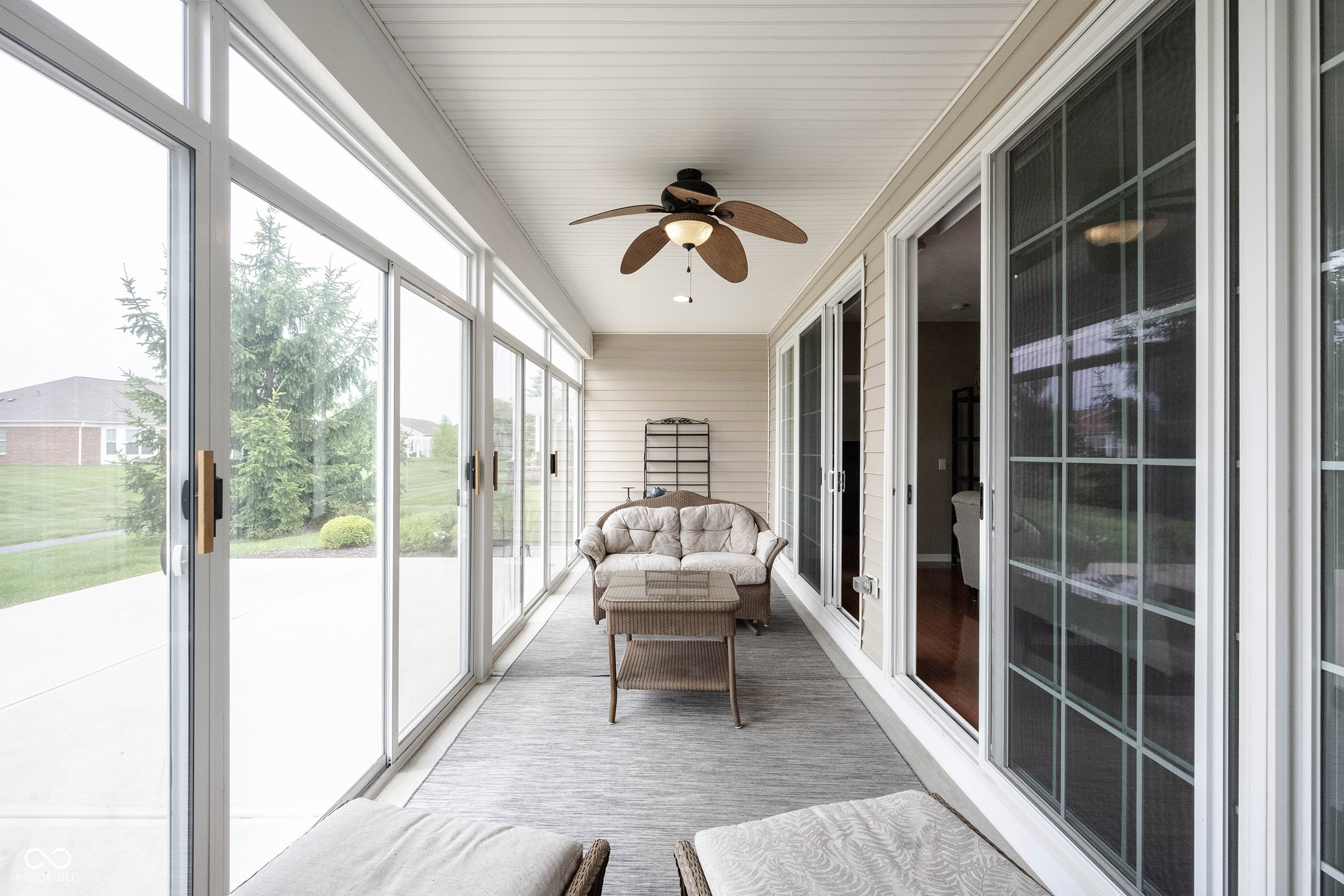 Enclosed sunroom with wicker seating, a ceiling fan, and large windows overlooking the lawn.