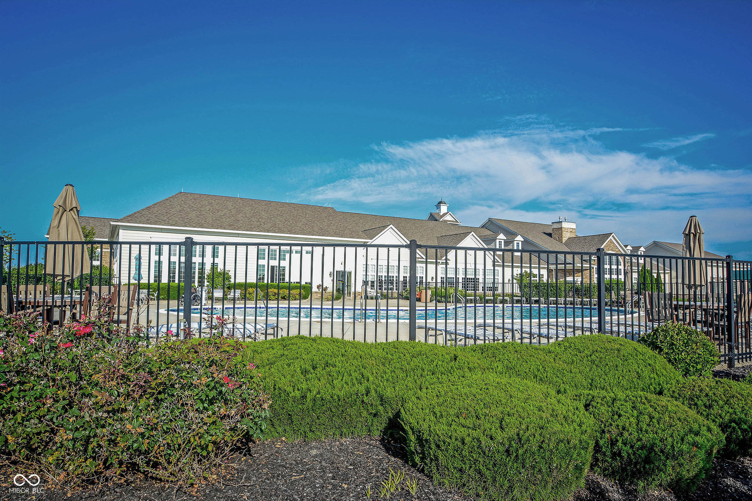 Outdoor pool in front of a white clubhouse, framed by hedges and a black metal fence.