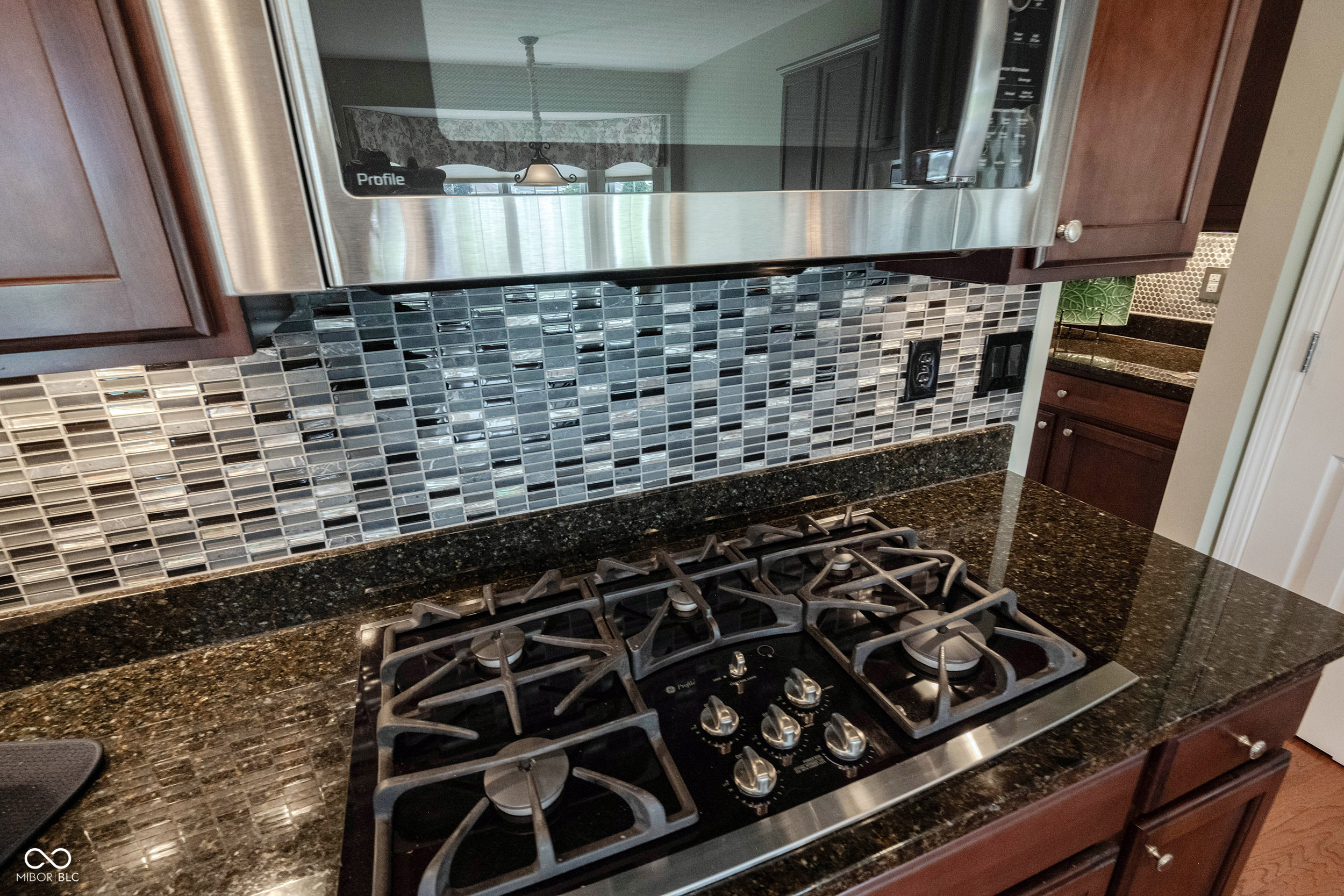 Kitchen cooktop beneath a stainless steel microwave, with dark granite counters and a gray glass-tile backsplash.