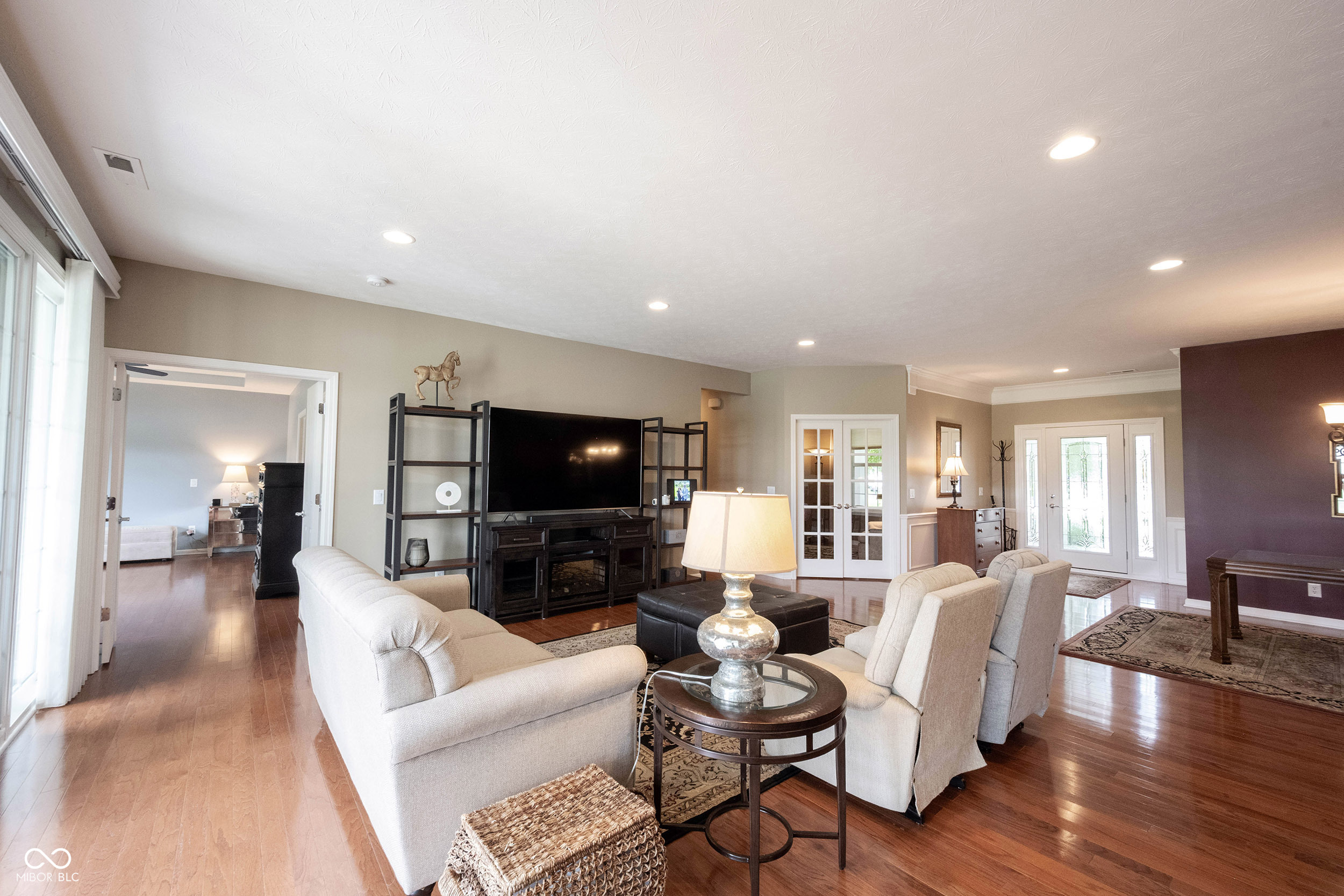 Furnished living room with hardwood floors, light seating, and a large television.