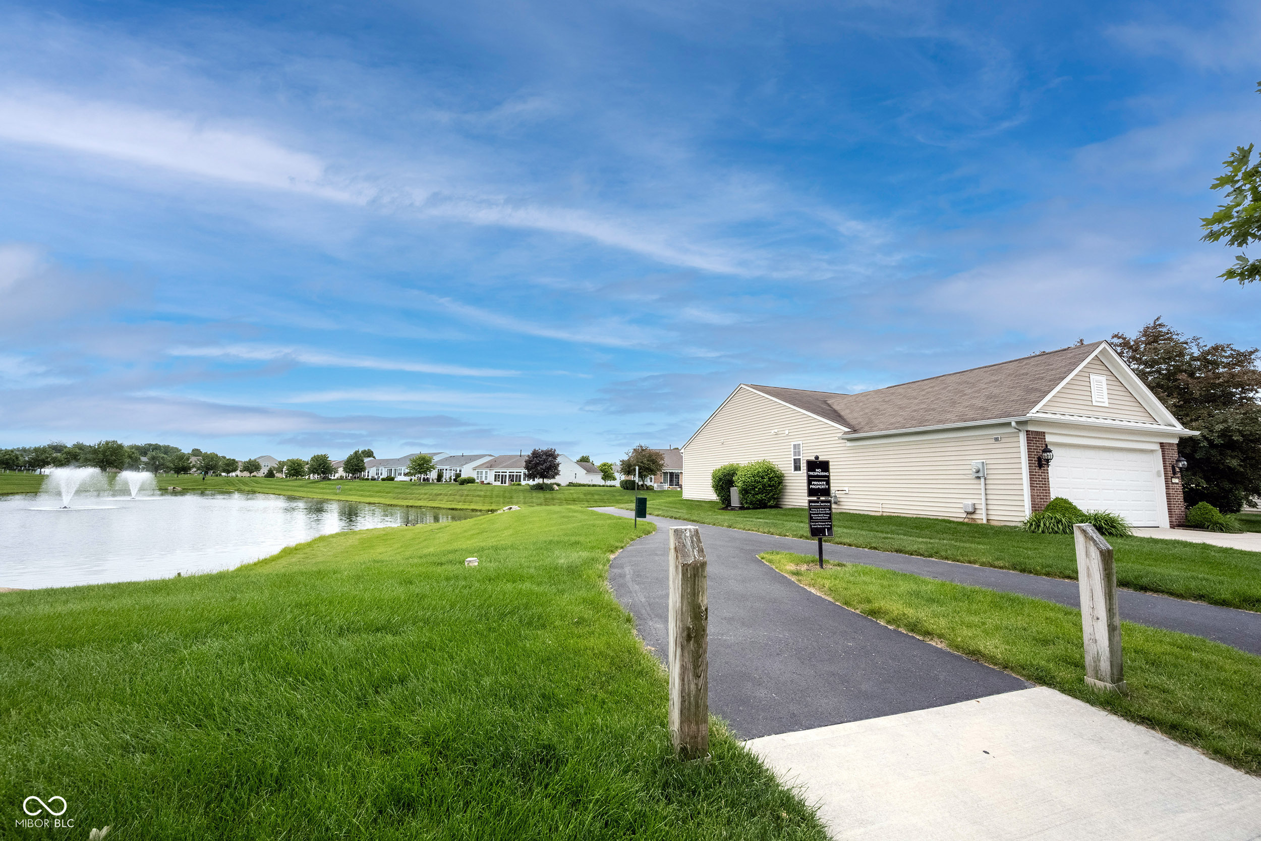 Exterior of a beige-sided home beside a pond, with a paved path and fountains.