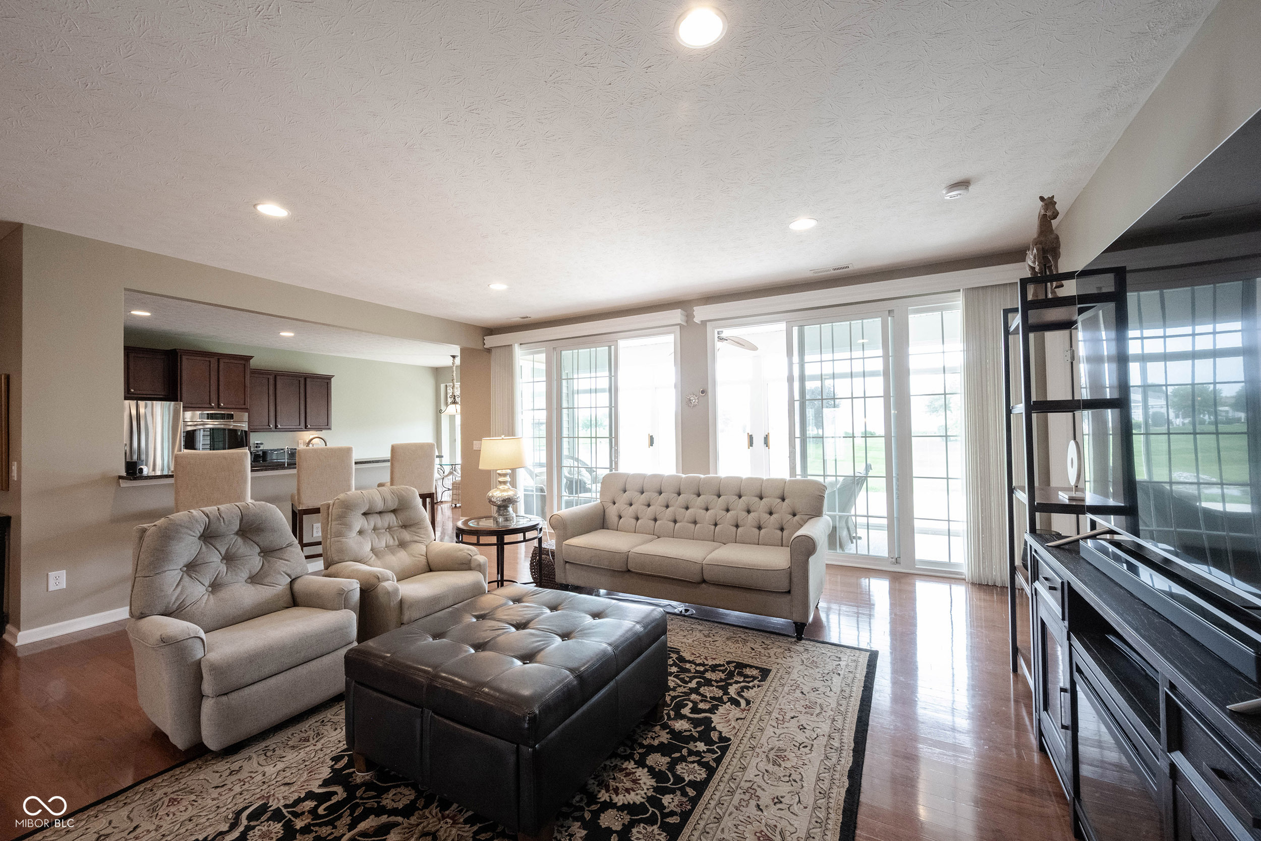 Furnished living room with beige seating, a leather ottoman, and large windows overlooking a lawn.