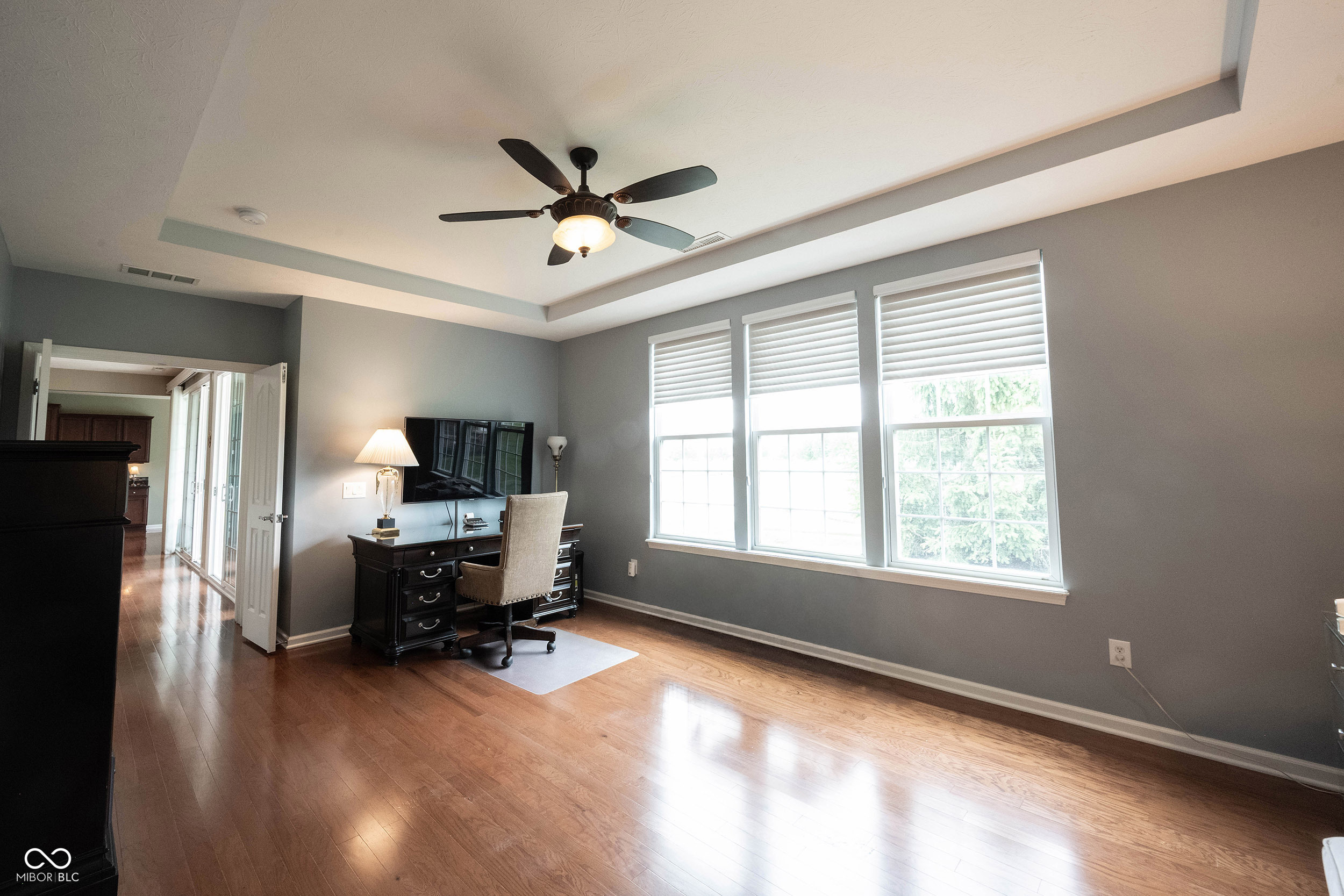 Home office with a dark desk, wood floors, and three tall windows with white blinds.