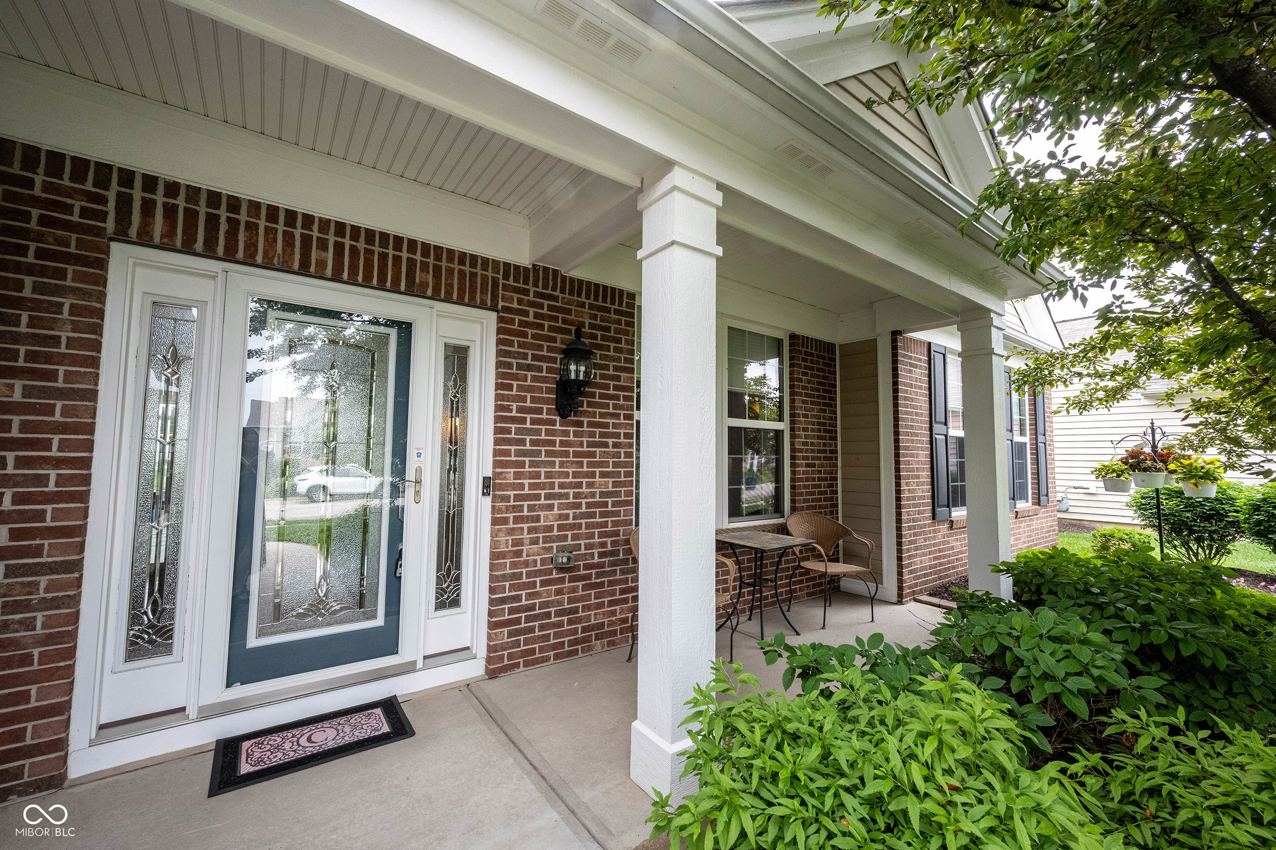 Brick home’s covered front porch with decorative glass entry door, white columns, and leafy landscaping.