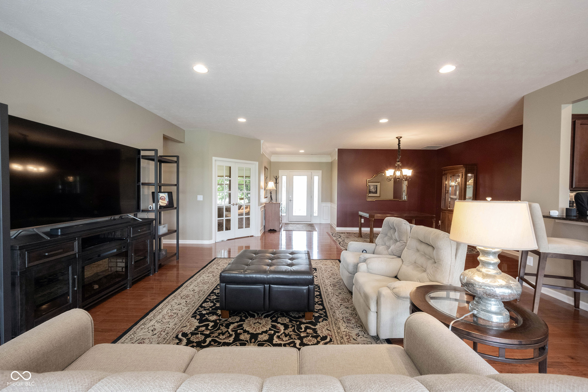 Furnished living room with hardwood floors, patterned rug, large television, and burgundy dining-room accent wall.