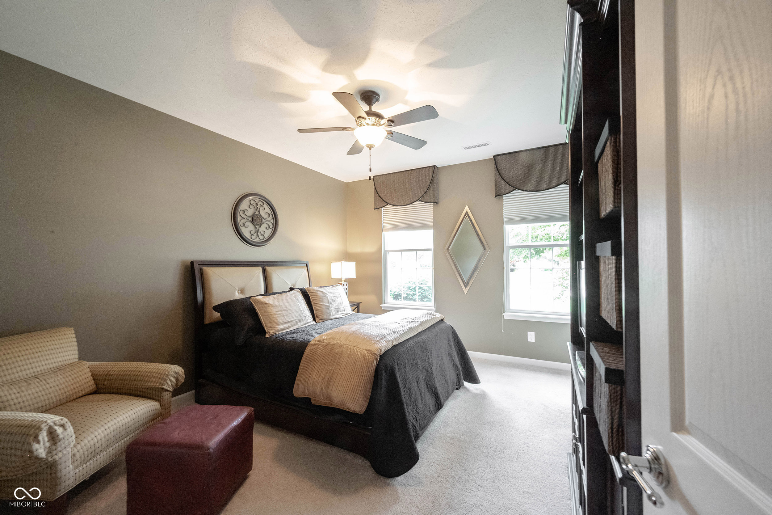 Bedroom with a dark bed, patterned armchair, two windows, and ceiling fan.