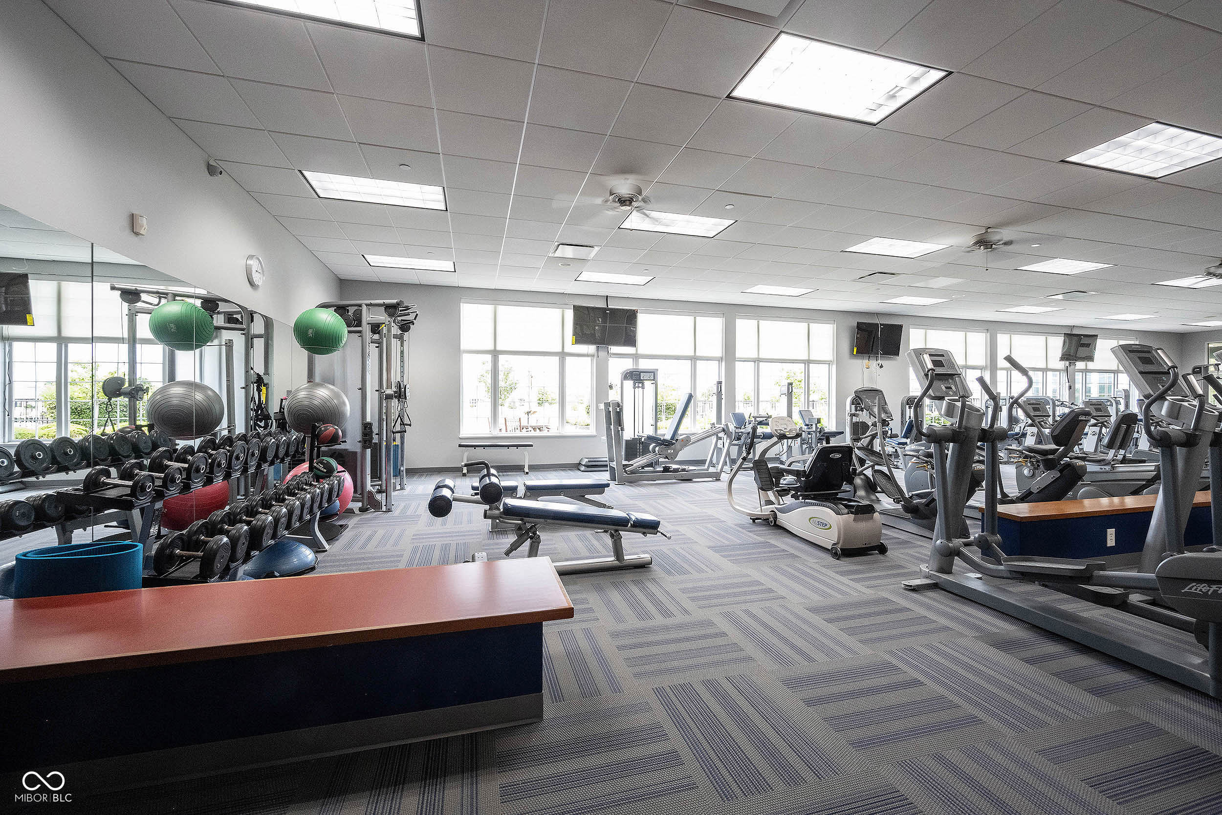 Fitness center with rows of weights, benches, and cardio machines beside large windows.