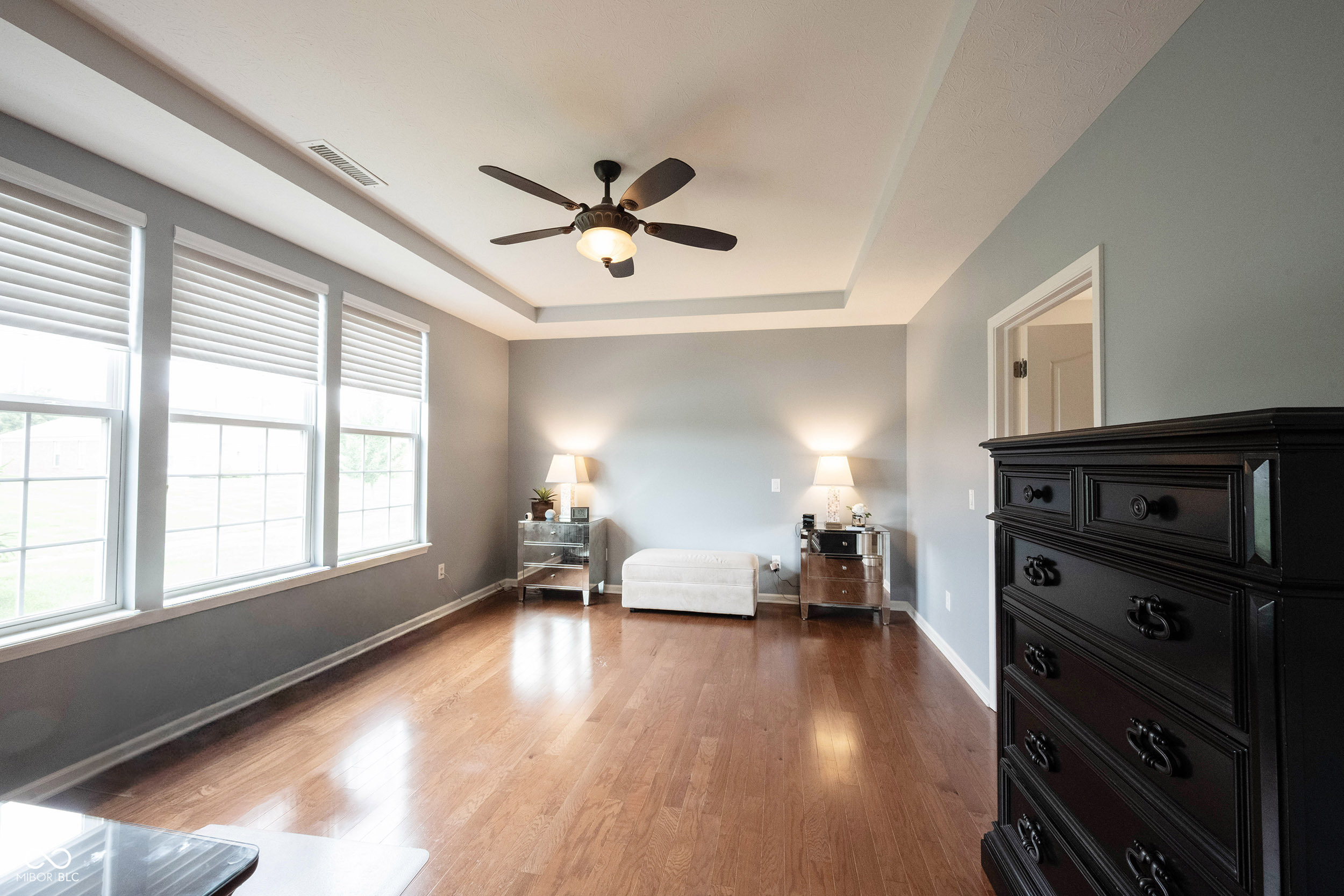 Bedroom with hardwood floors, three tall windows, a ceiling fan, mirrored nightstands, and a large dark dresser.