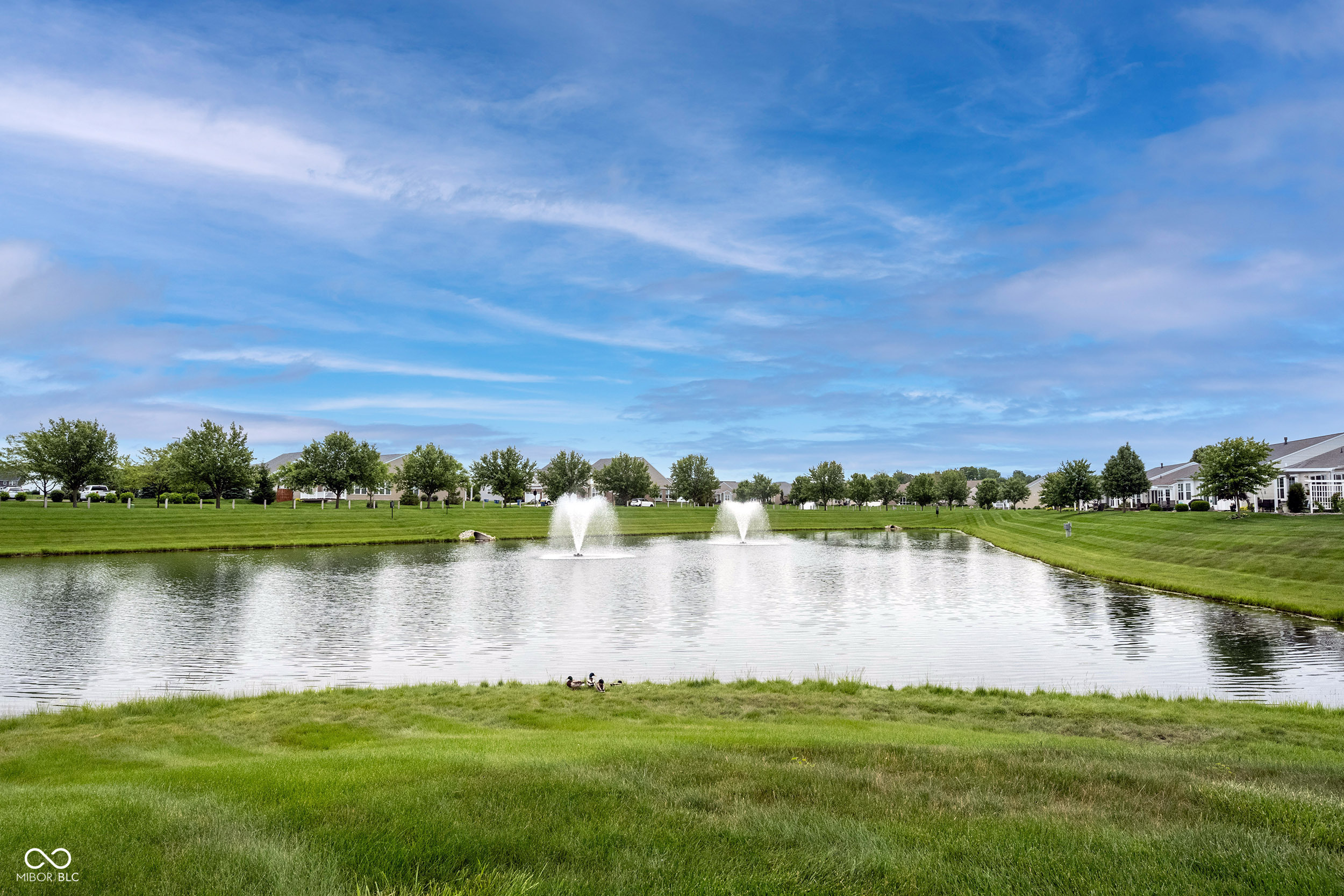 Neighborhood pond with two fountains, grassy banks, and homes along the far shore.