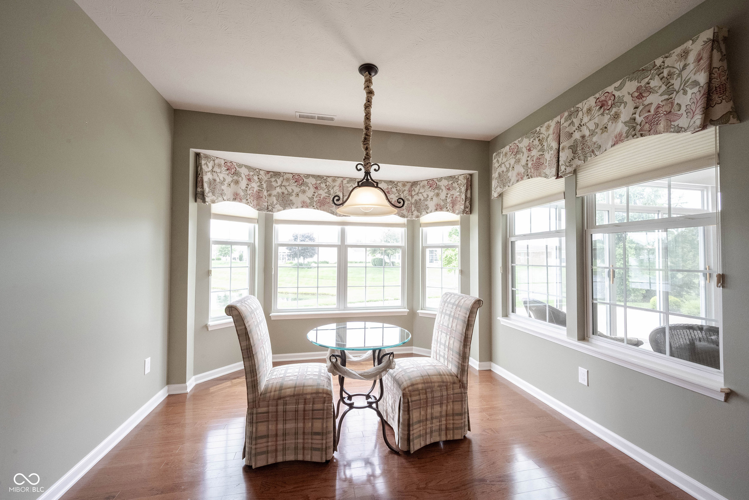 Breakfast nook with a round glass table, two plaid chairs, and large windows with floral valances.
