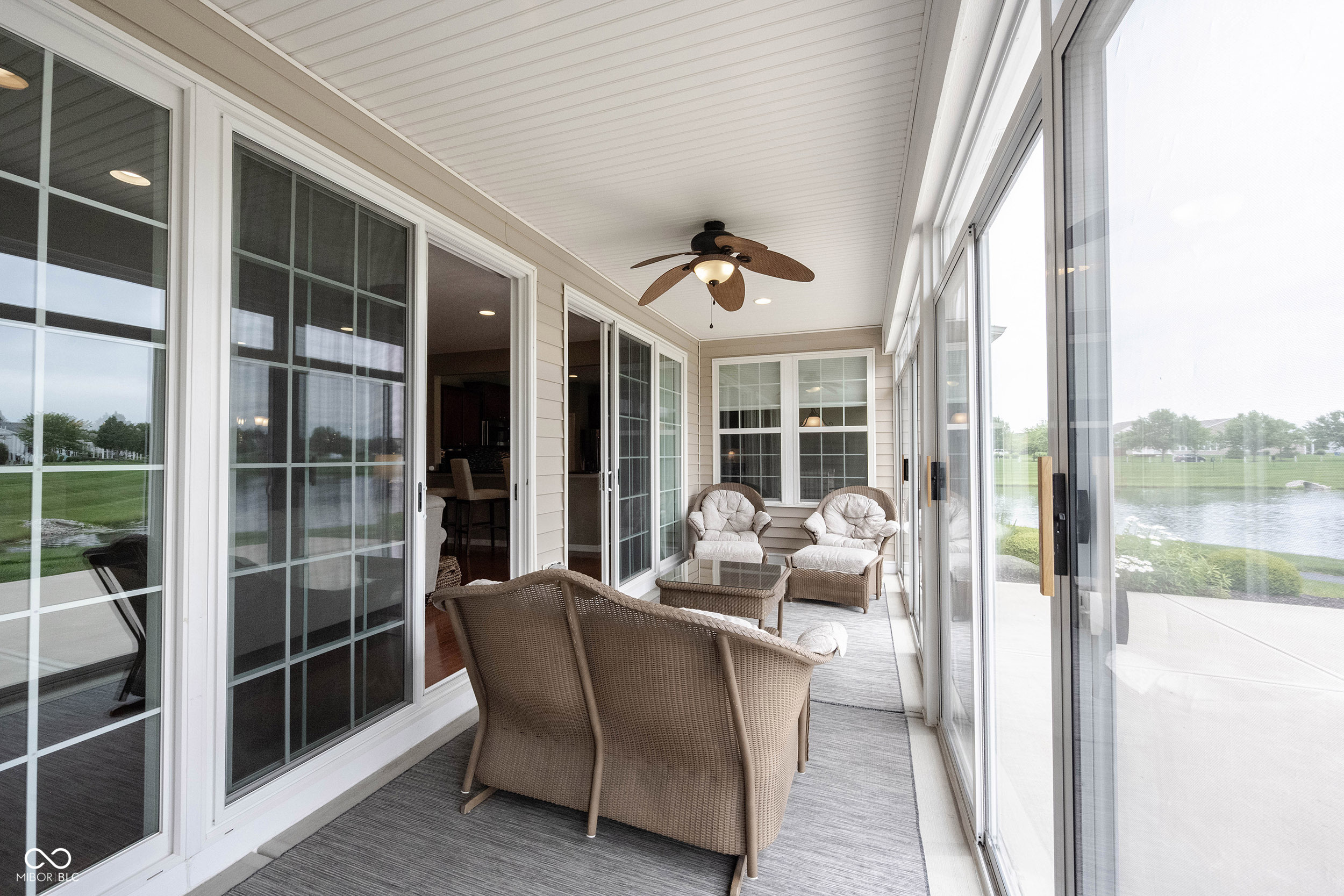 Glass-enclosed sunroom with wicker seating and a pond view through the windows.