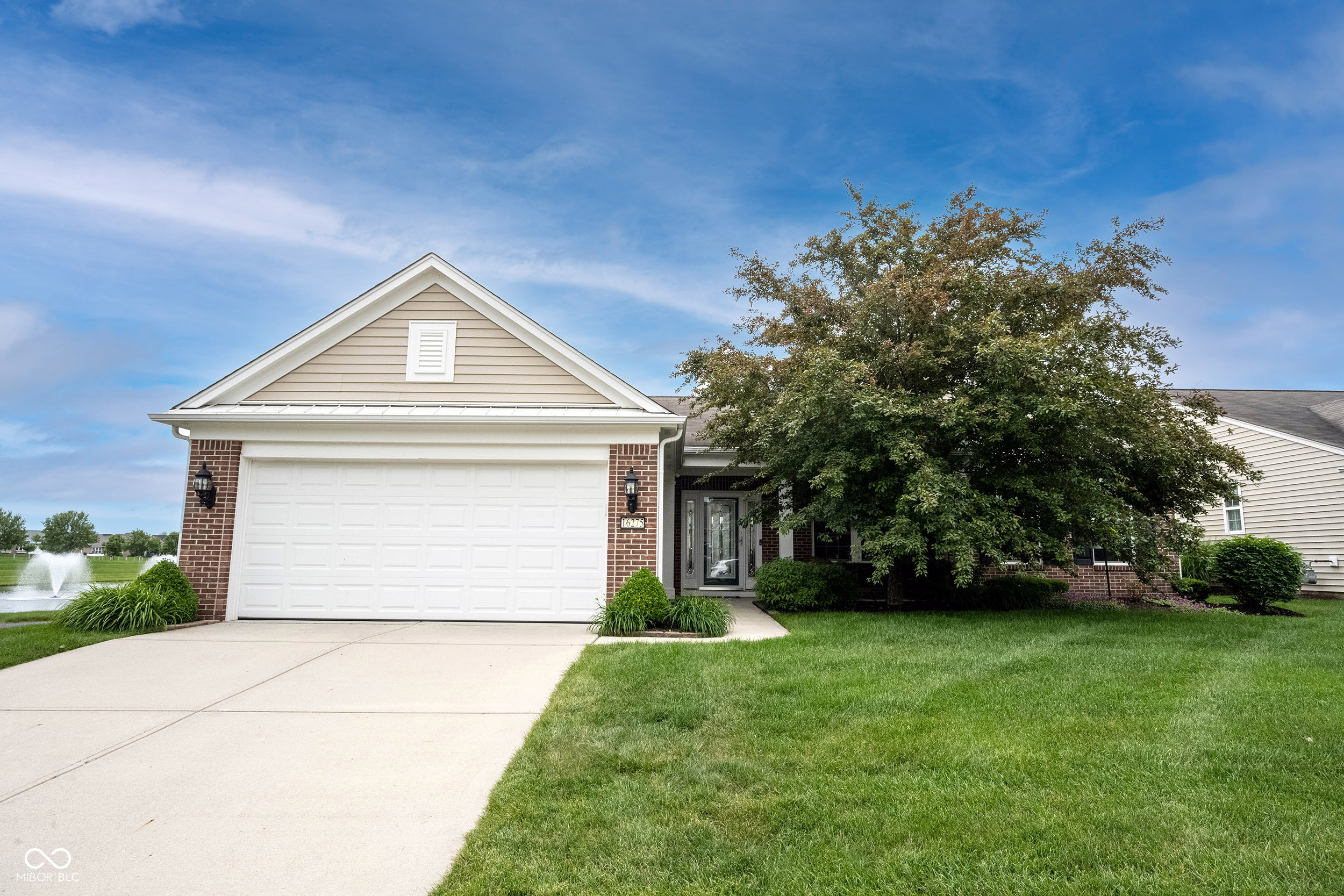 House exterior with a white garage, mature tree, broad driveway, and pond fountain nearby.