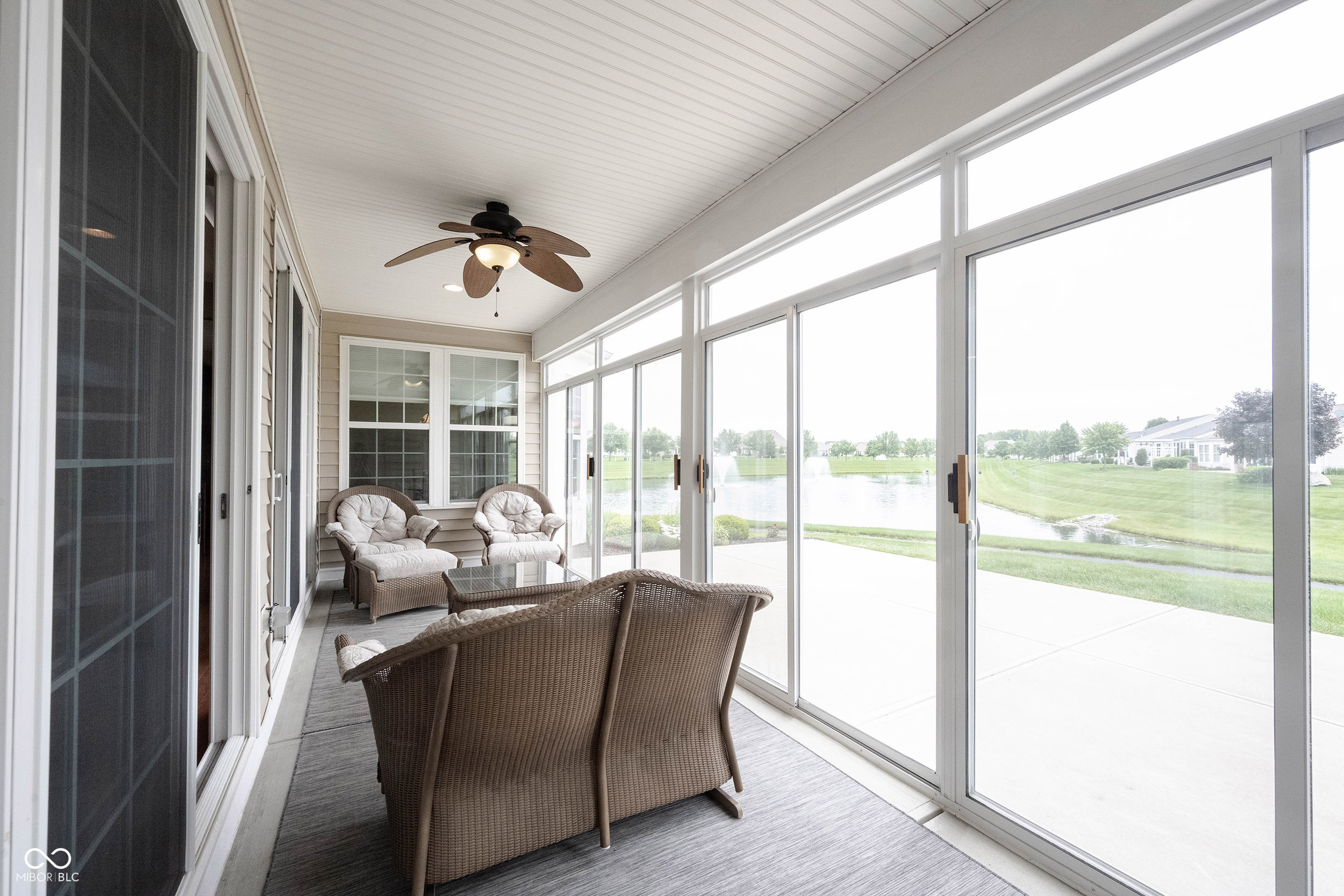 Furnished sunroom with wicker seating and wide windows overlooking a pond and landscaped lawn.