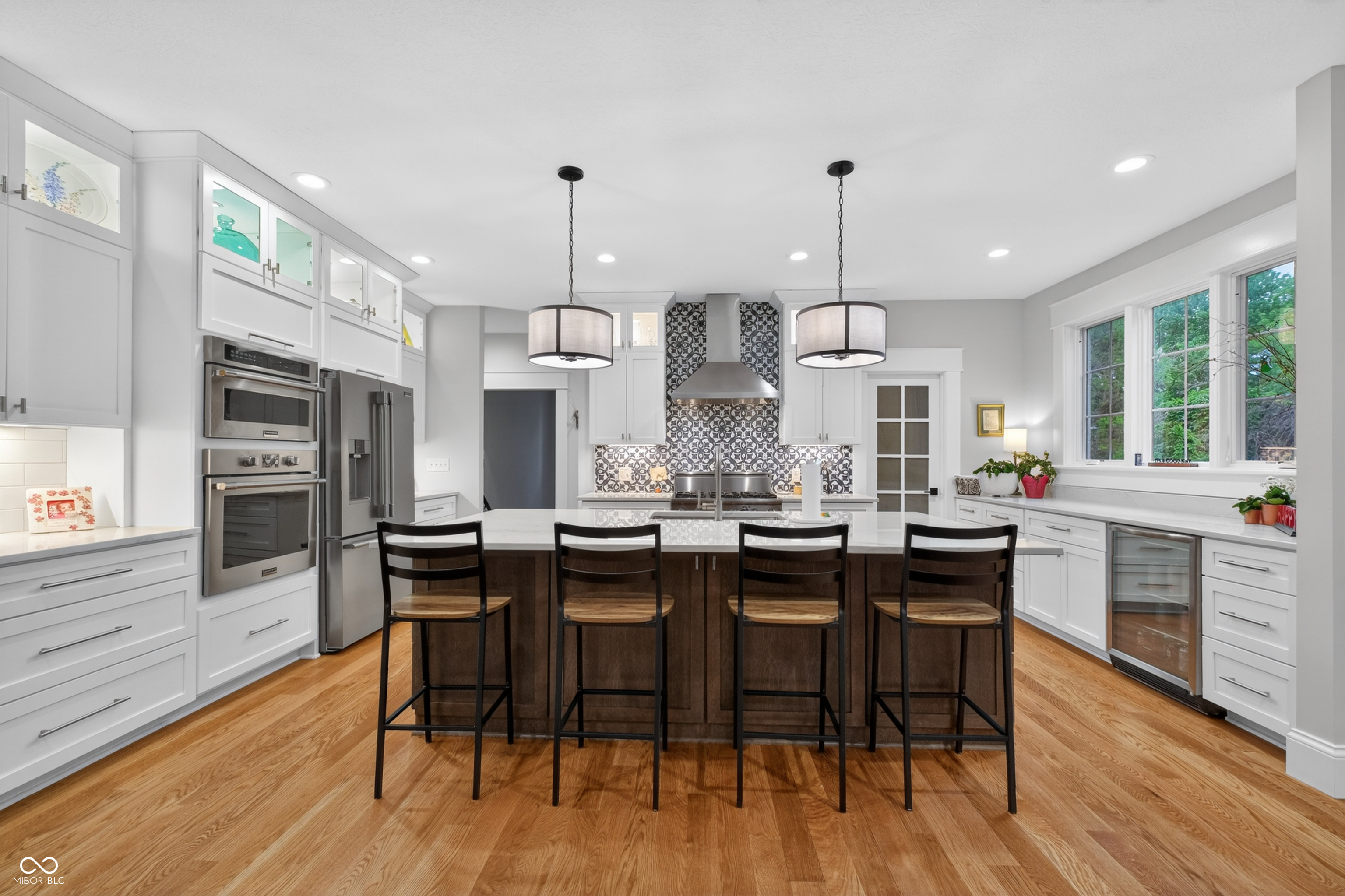 Bright kitchen with a dark wood island, four stools, white cabinets, and large windows overlooking greenery.