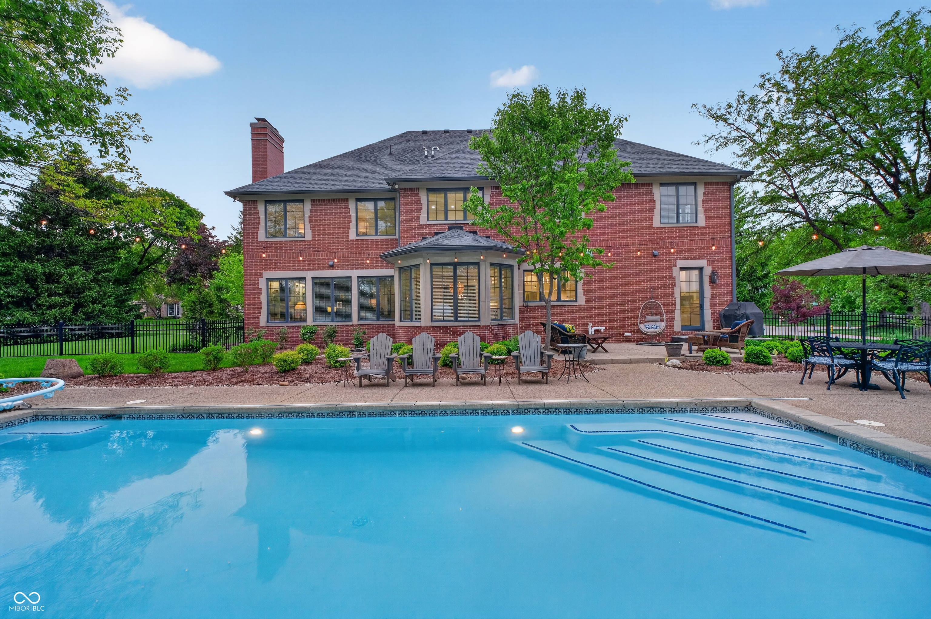 Red brick home behind a swimming pool, with a furnished patio and string lights at dusk.