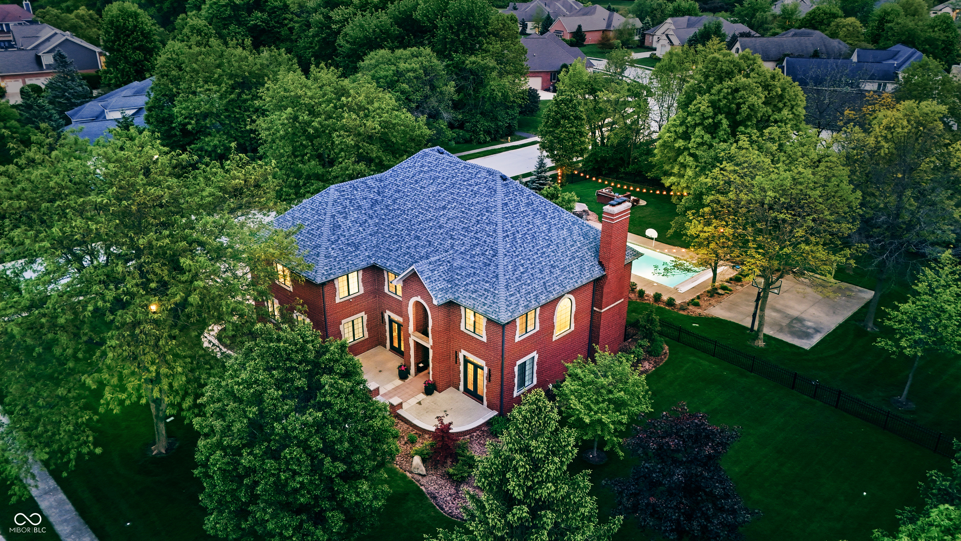 Aerial view of a red-brick home with a fenced pool, patio, and mature trees.