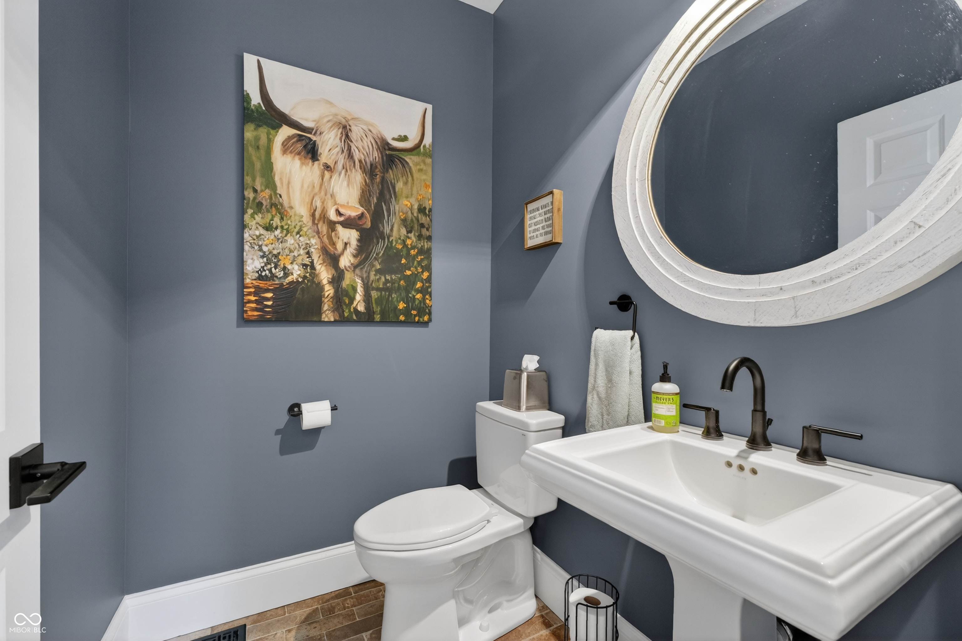 Bathroom with slate-blue walls, a white pedestal sink, round mirror, and Highland cow painting.