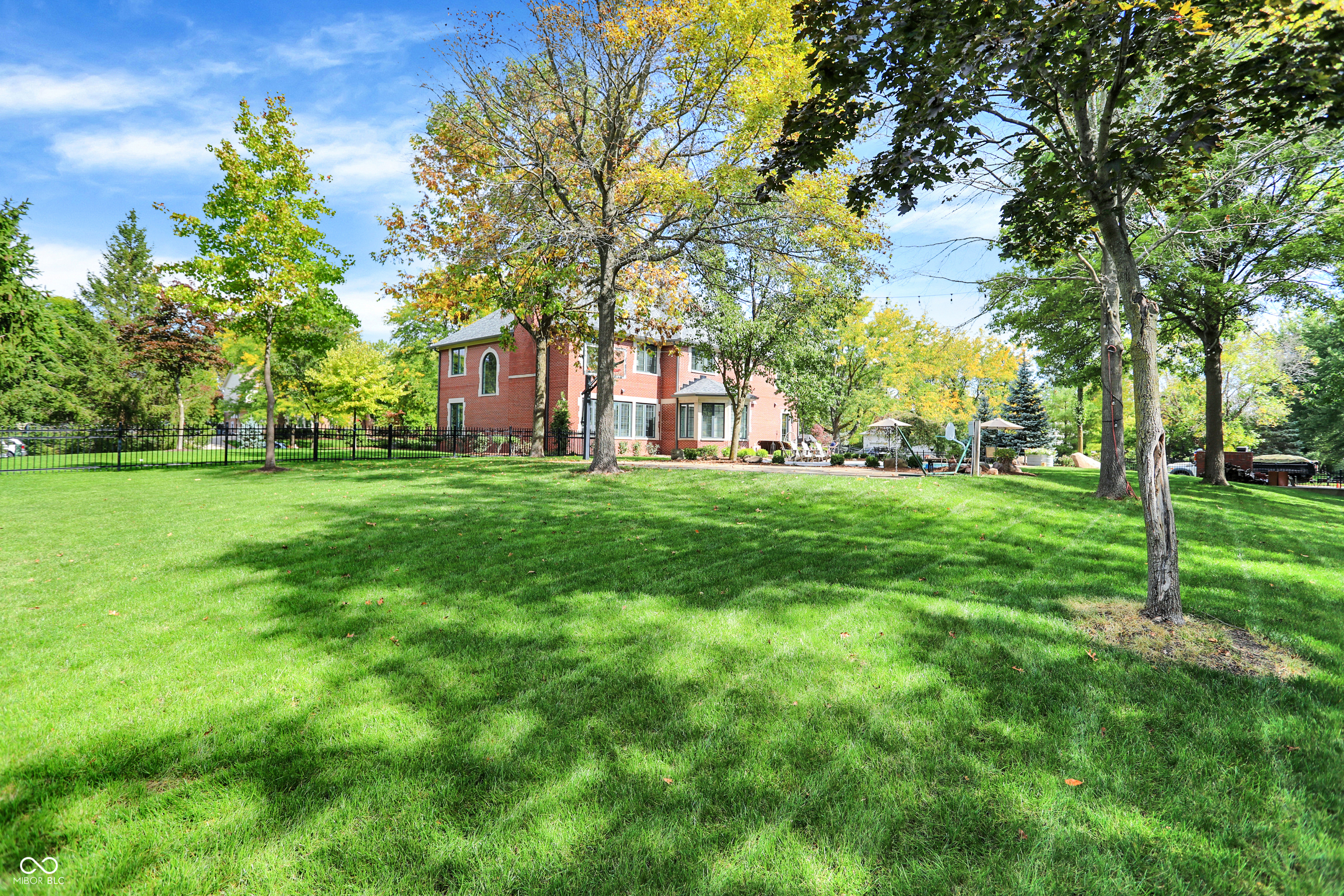 Red-brick two-story house beyond a broad lawn, with mature trees, patio seating, and a backyard pool.