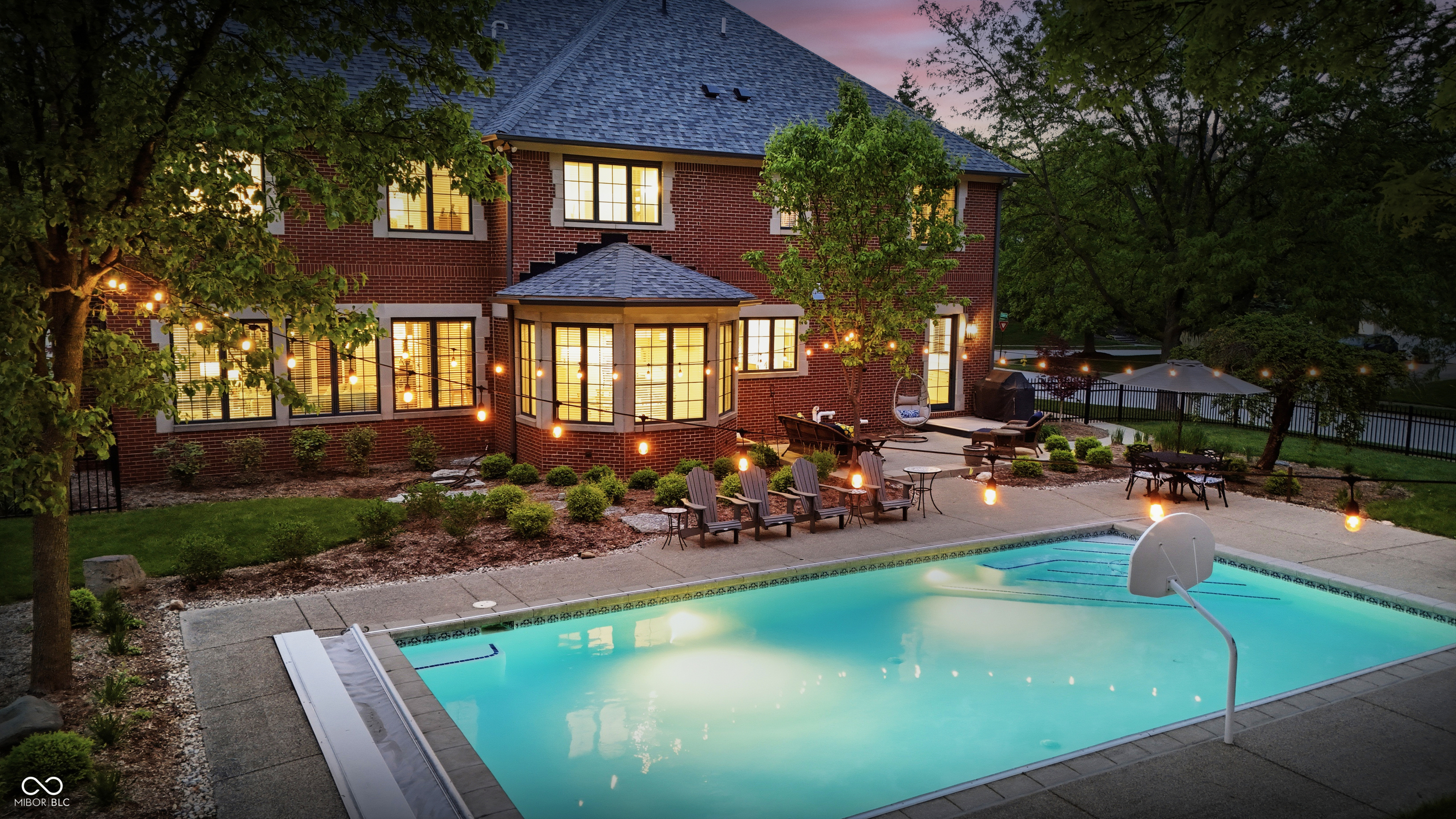 Red-brick home and illuminated patio beside a backyard swimming pool at dusk.