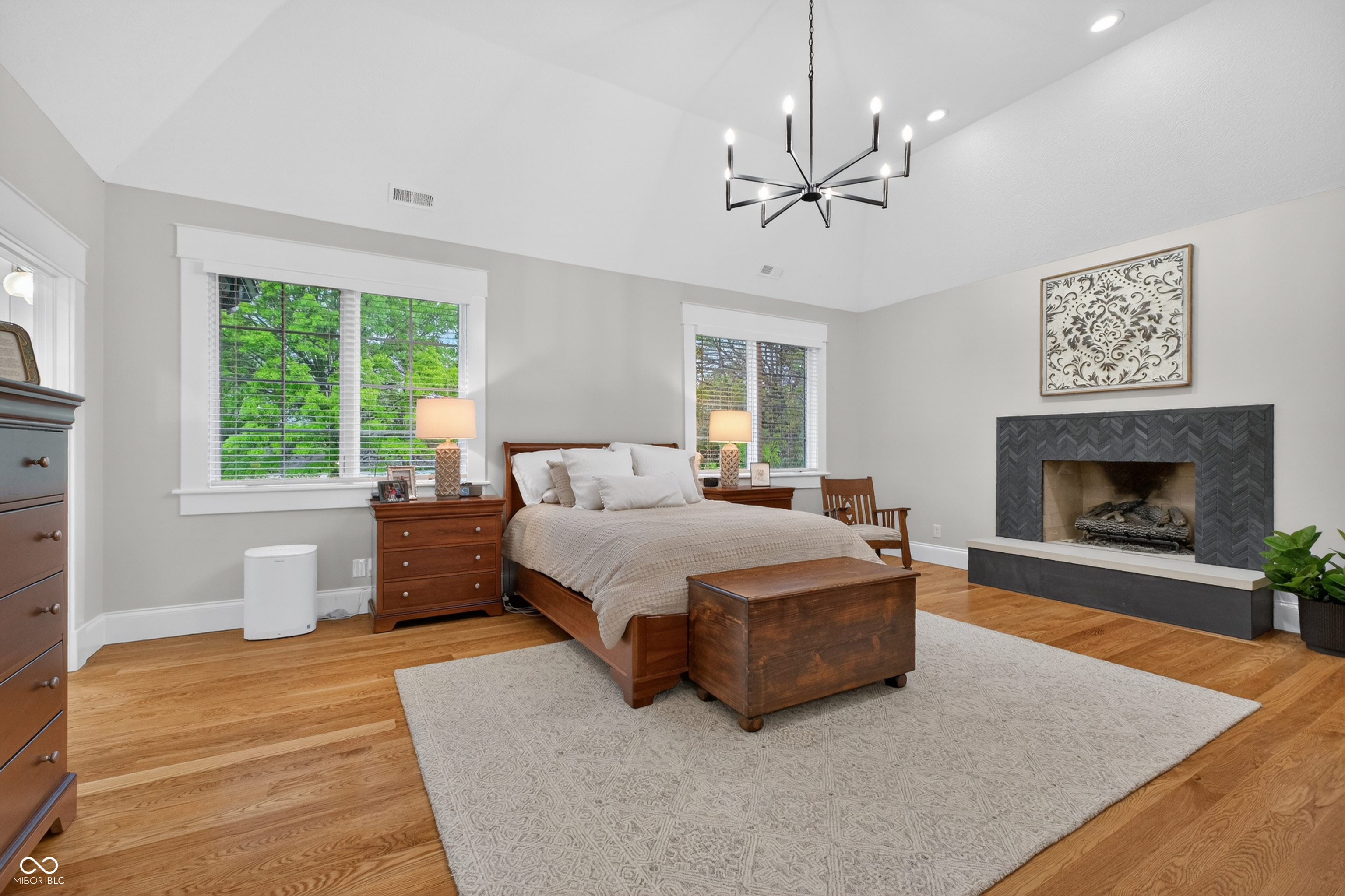 Furnished bedroom with a vaulted ceiling, hardwood floors, and a fireplace beside the bed.