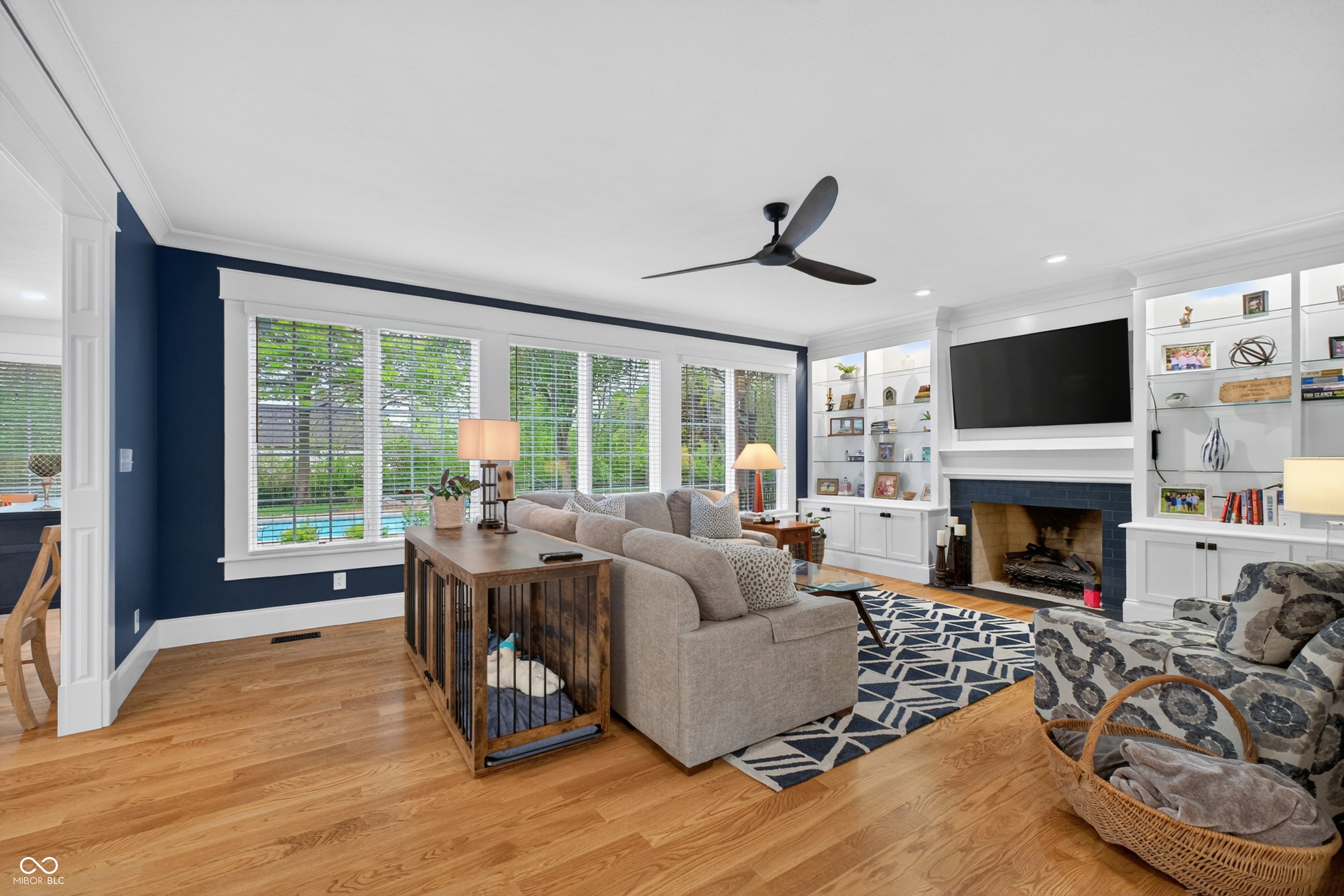 Living room with built-in shelves, fireplace, hardwood floors, and windows overlooking a backyard pool.