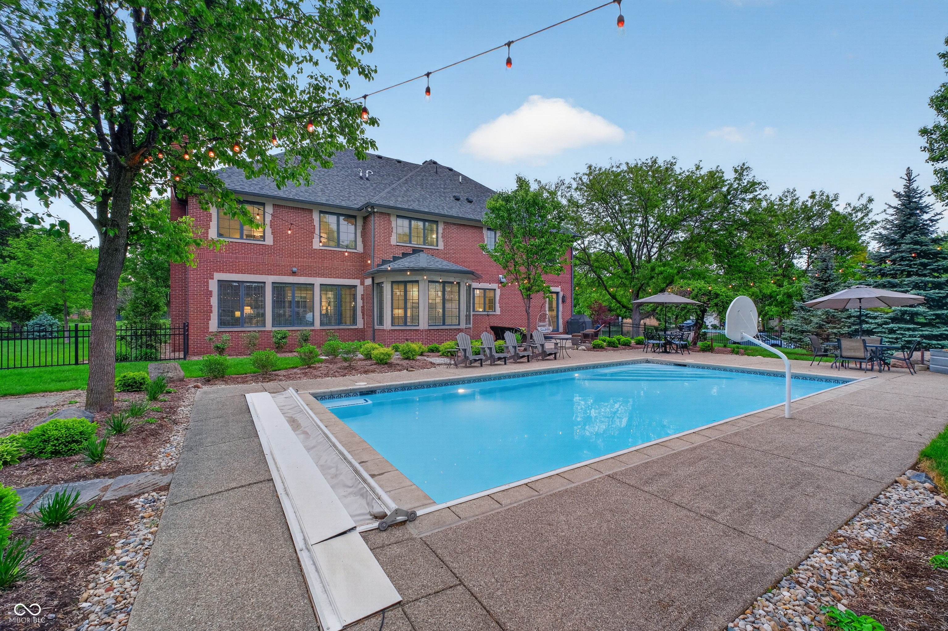 Backyard pool behind a red brick home, with patio seating, string lights, and a basketball hoop.