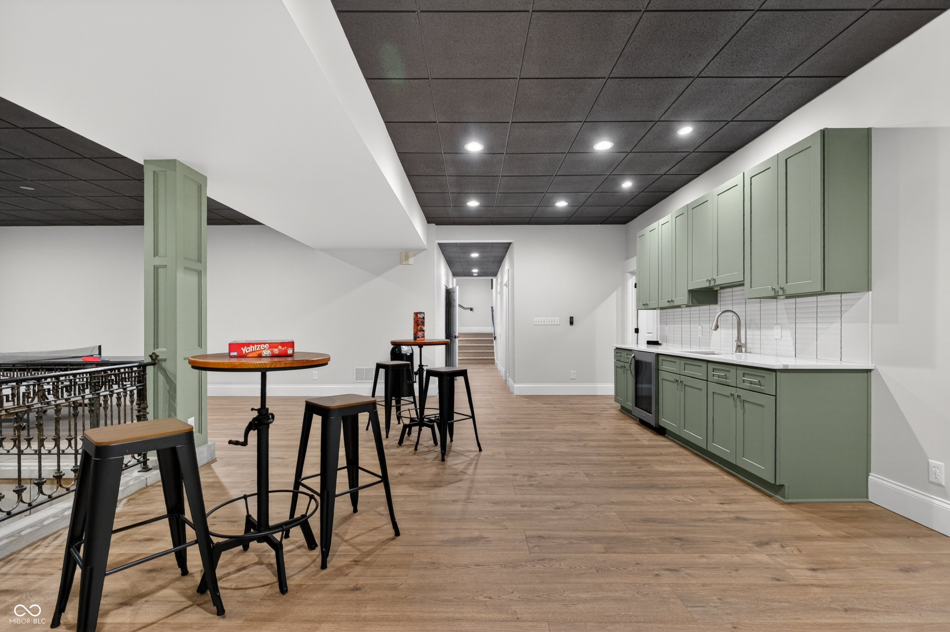 Lower-level recreation room with a sage-green kitchenette, bar stools, and wood-look flooring.