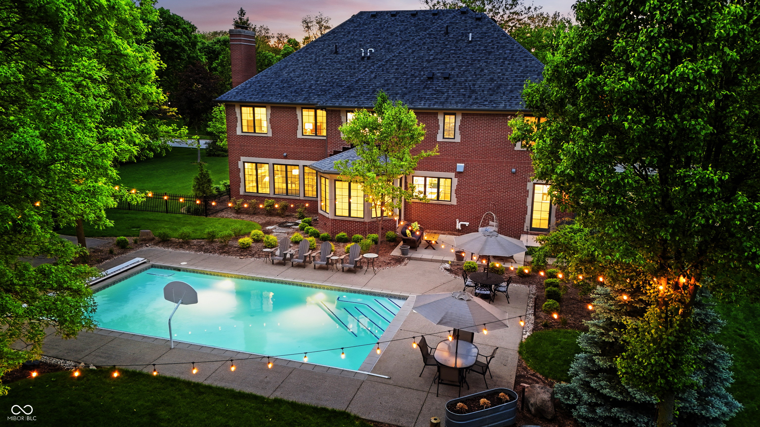 Red-brick home and backyard pool lit at dusk, with patio seating and string lights.