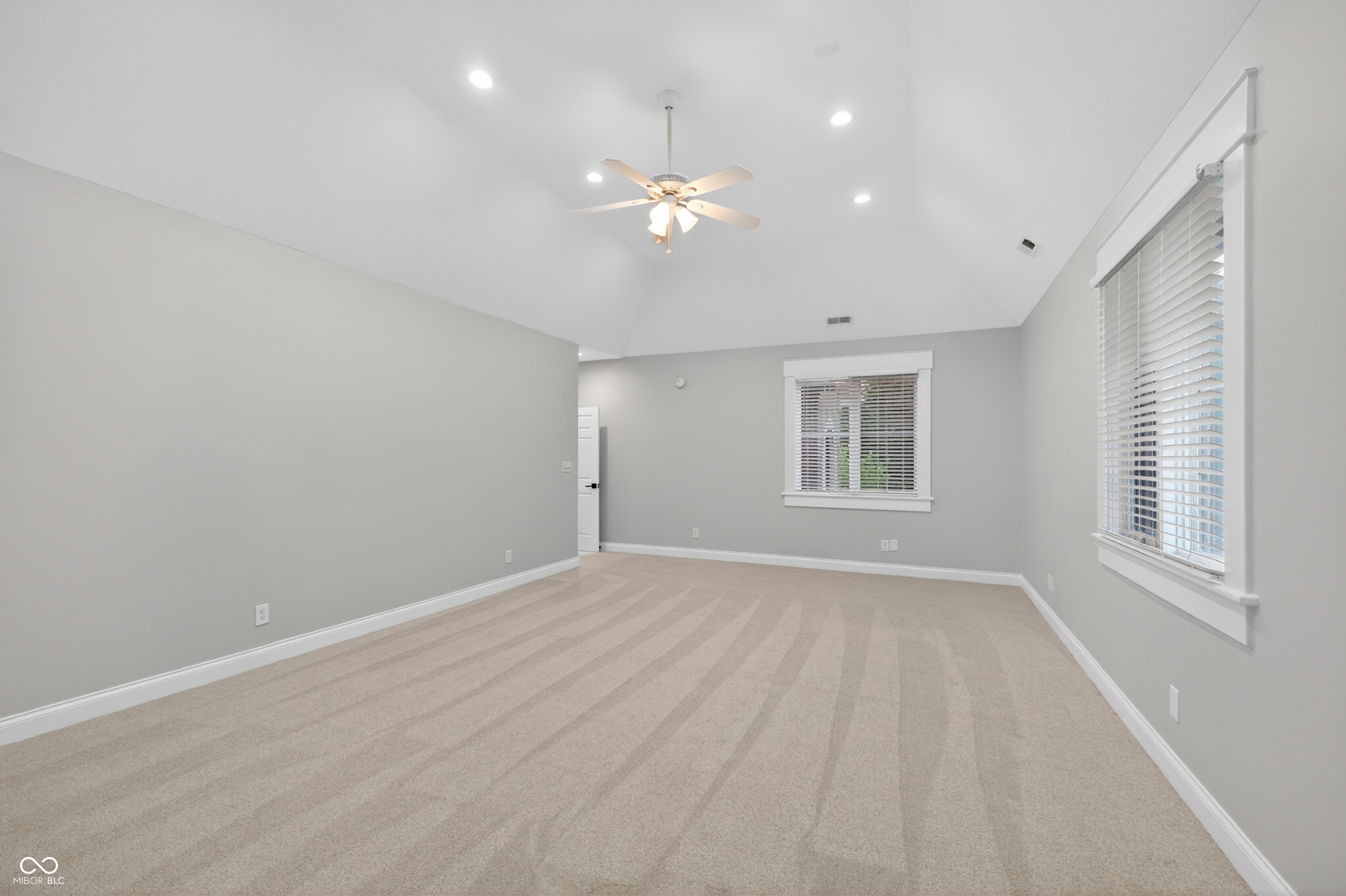 Empty carpeted room with a high ceiling, ceiling fan, and two windows with white blinds.