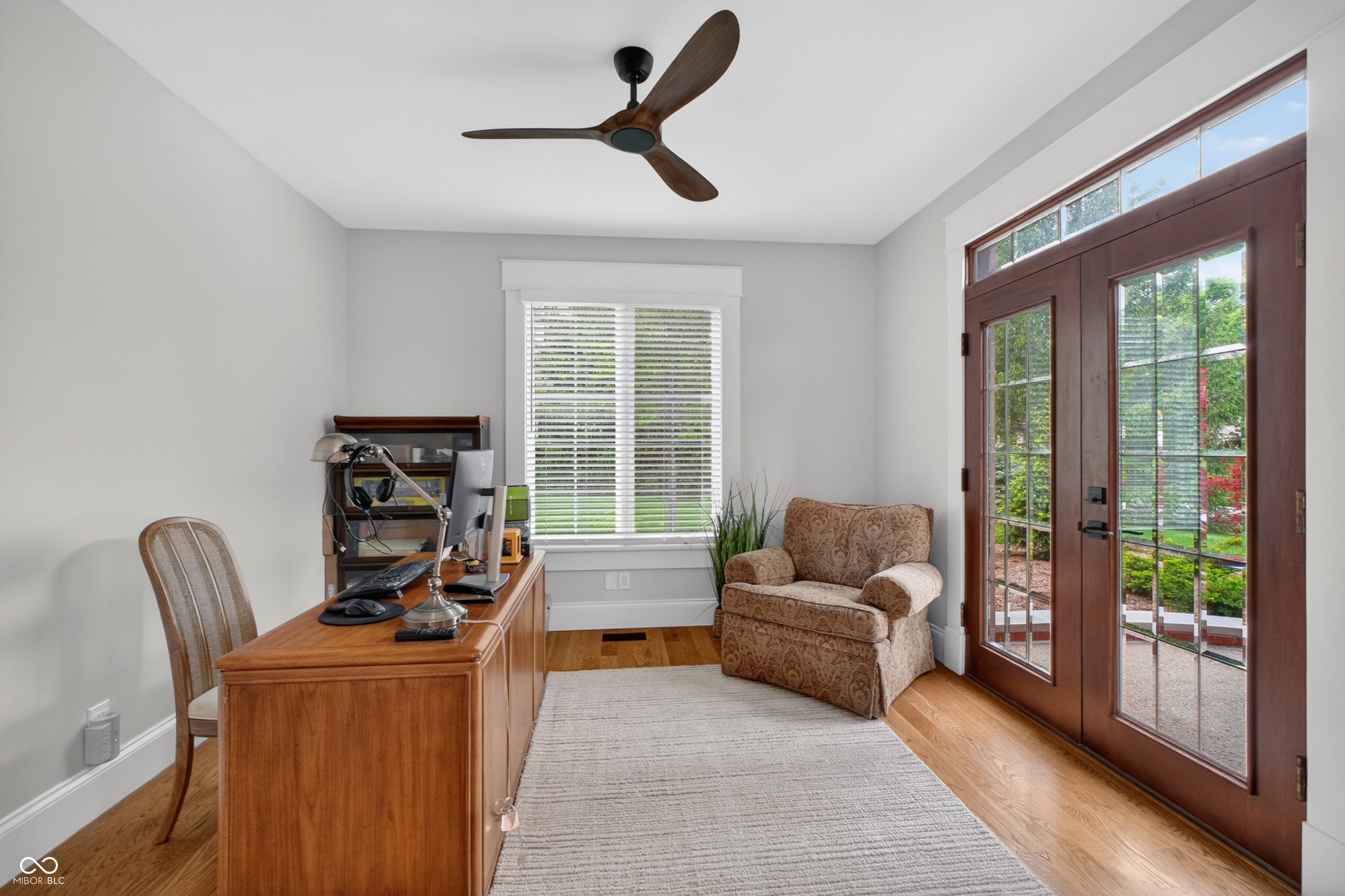 Home office with a wooden desk, patterned armchair, and dark wood-framed glass doors.