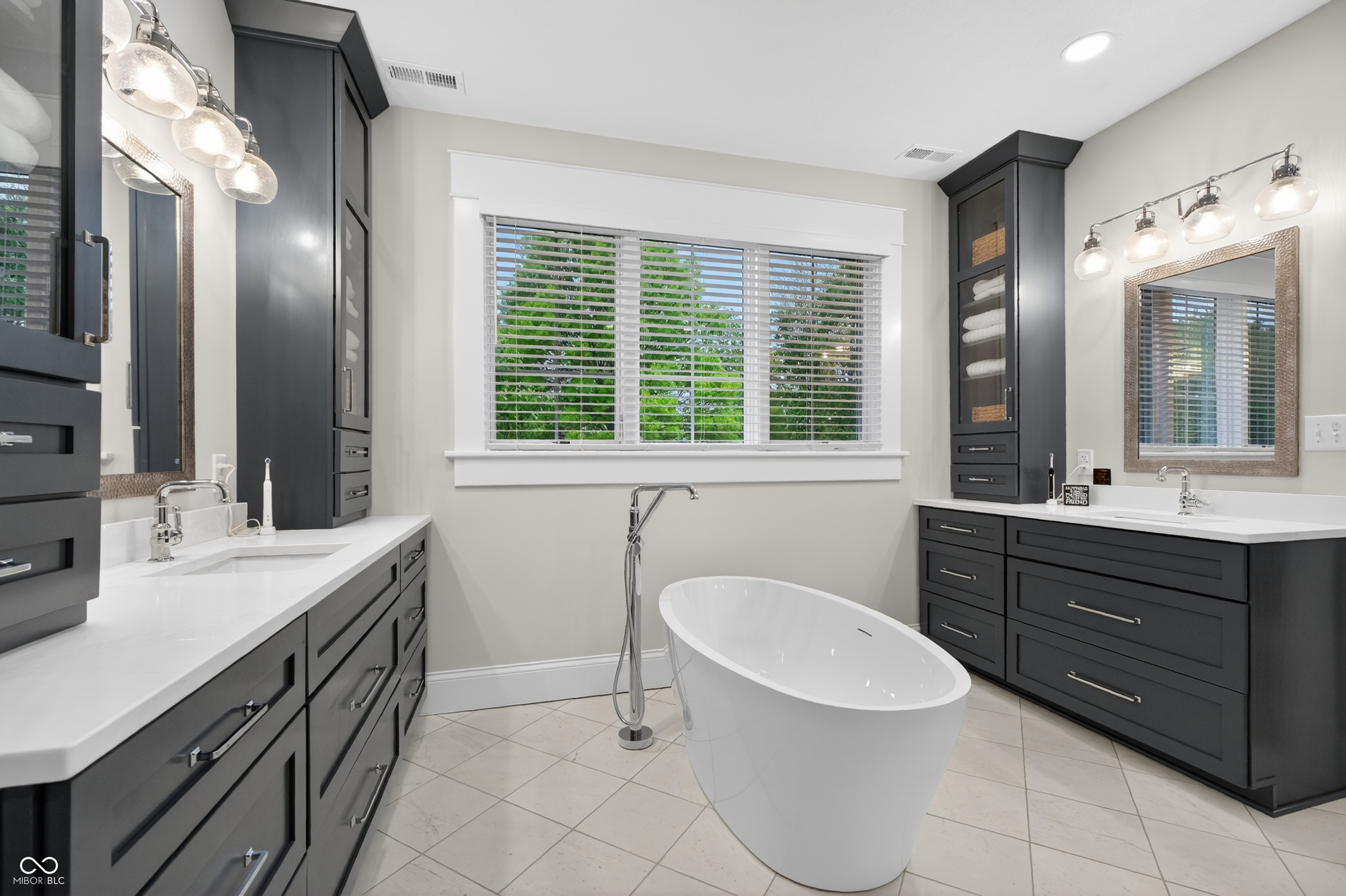 Bathroom with a freestanding soaking tub, charcoal vanities, and a wide window with white blinds.