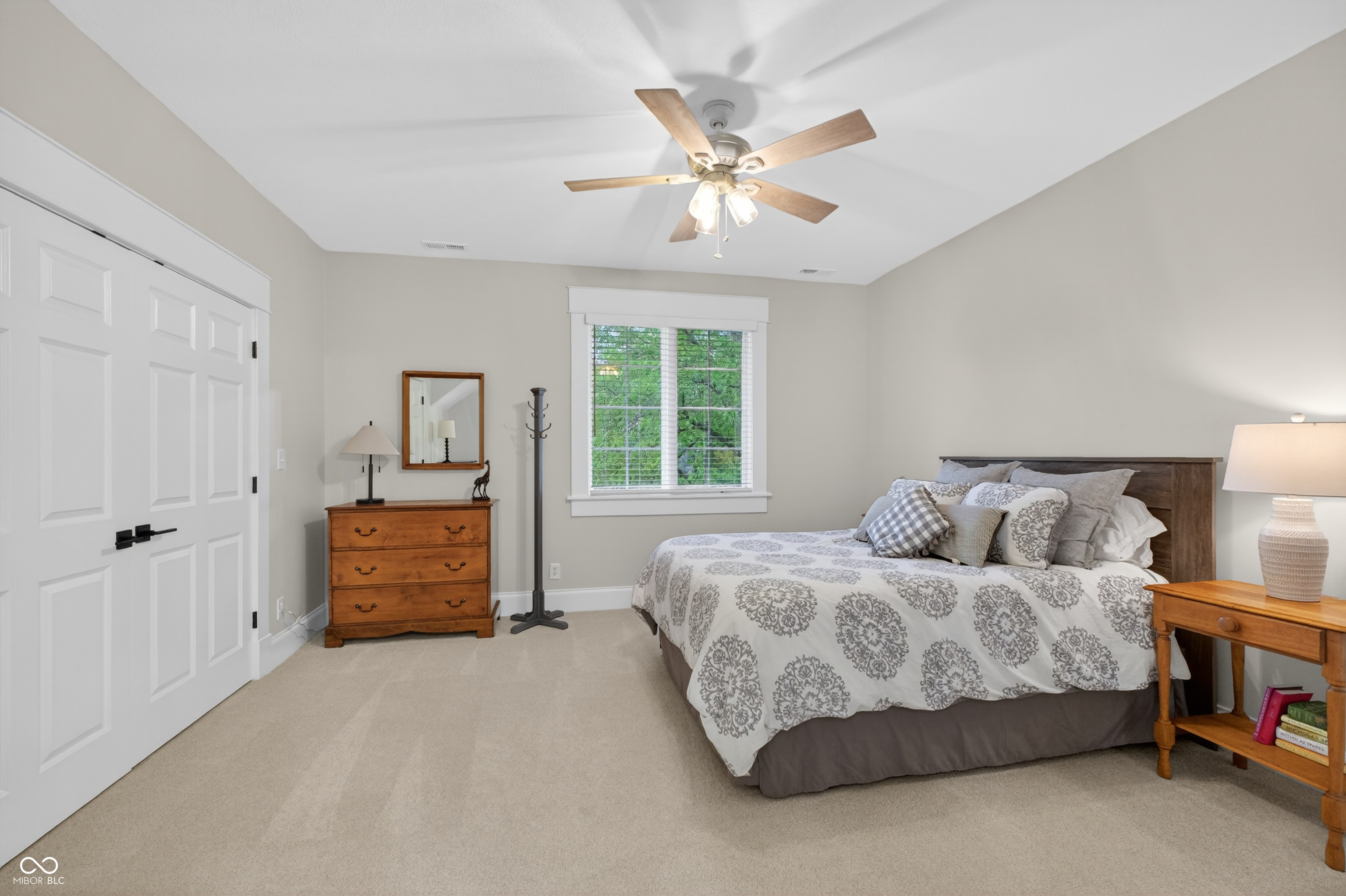 Furnished bedroom with a patterned bedspread, white closet doors, and a window overlooking green trees.