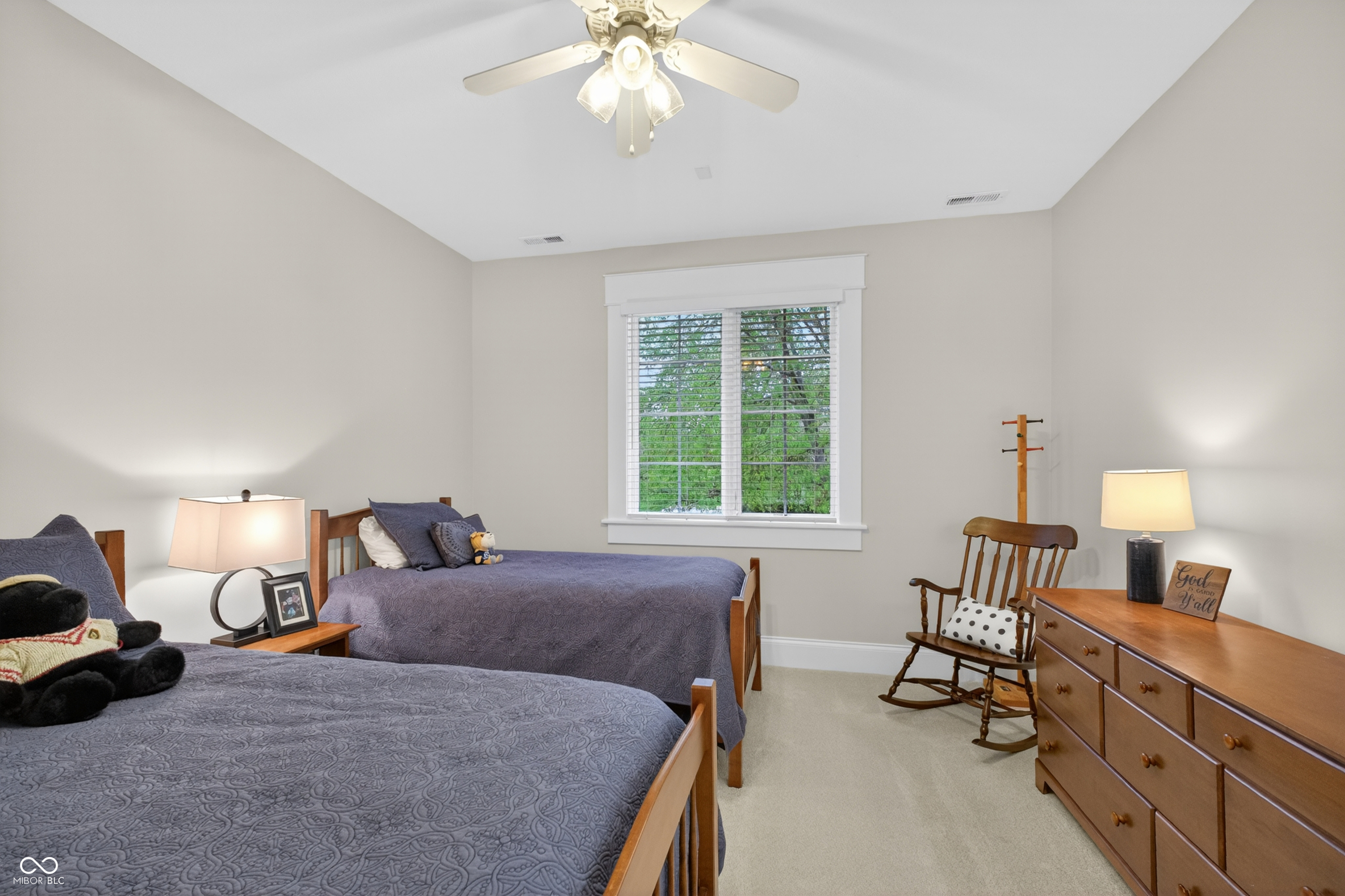 Furnished bedroom with two twin beds, a window with white blinds, and a wooden dresser.
