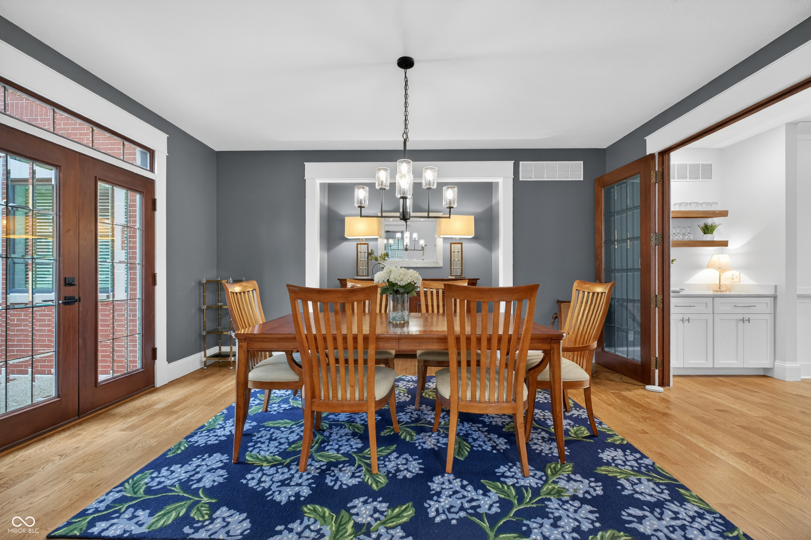Dining room with a wooden table, blue floral rug, chandelier, and wood-framed glass doors.