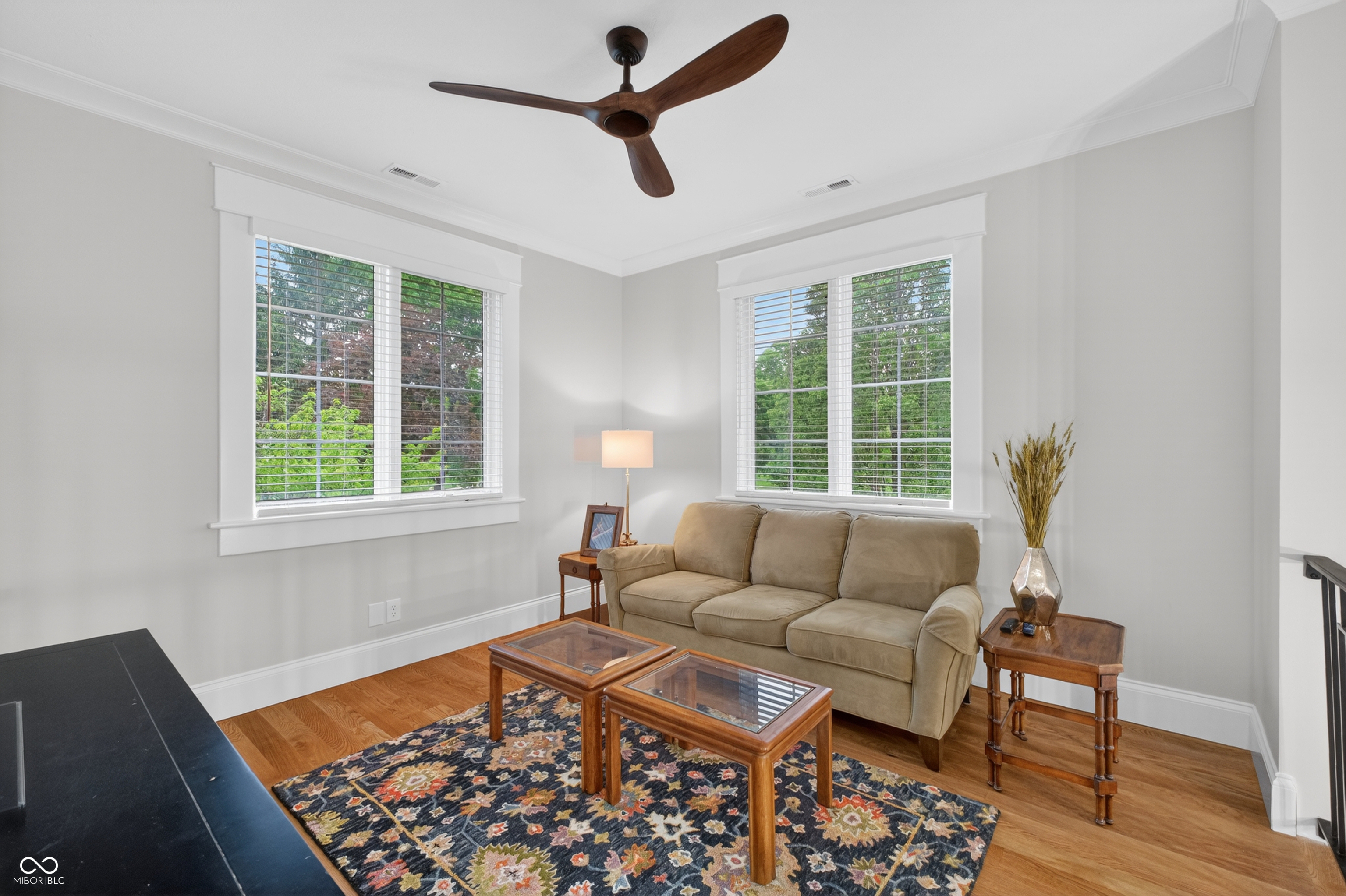 Staged living room with a tan sofa, two large windows, hardwood floors, and a patterned rug.