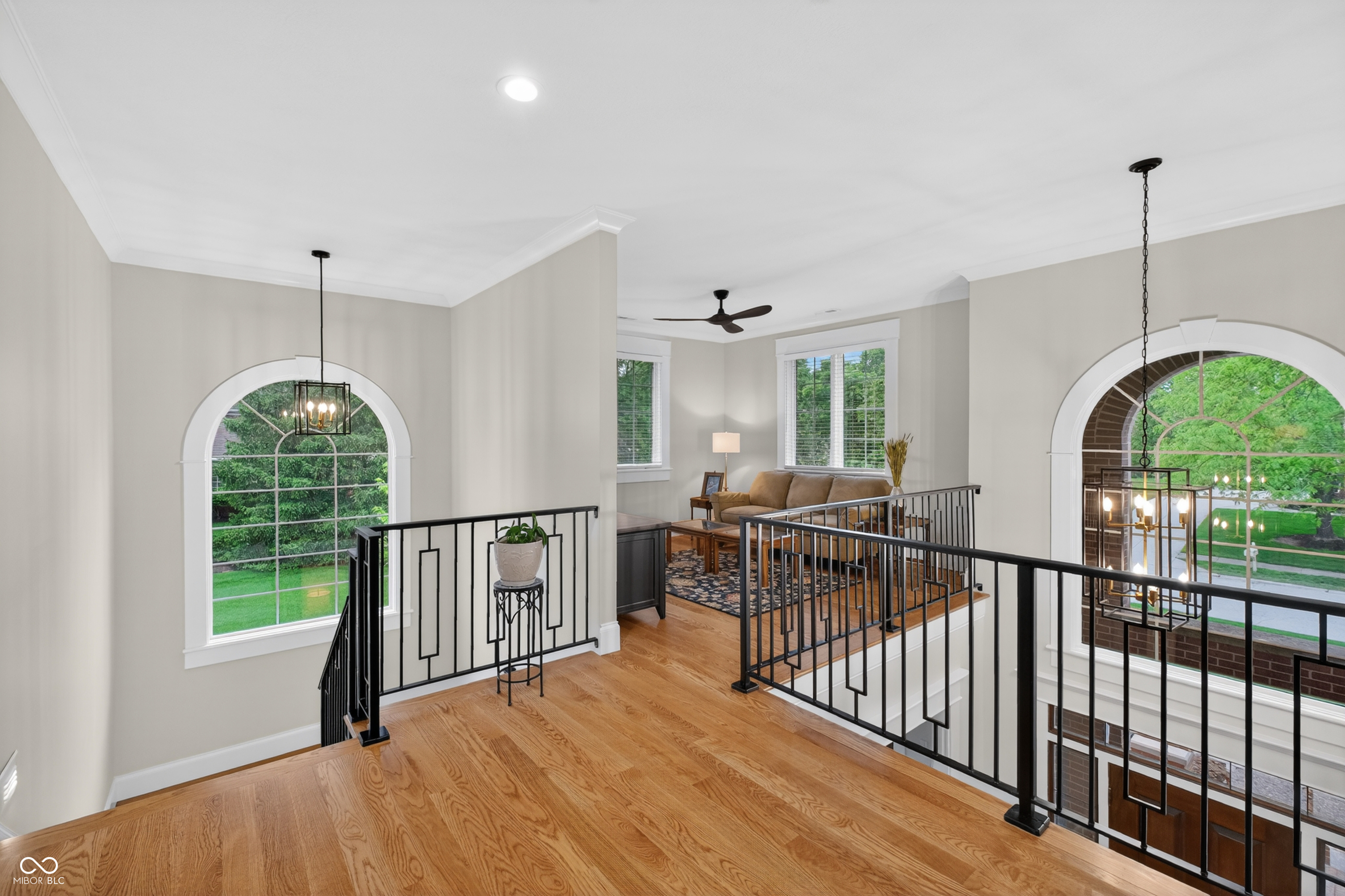 Upstairs landing with hardwood floors, black railings, arched windows, and a tan sofa in the living room beyond.