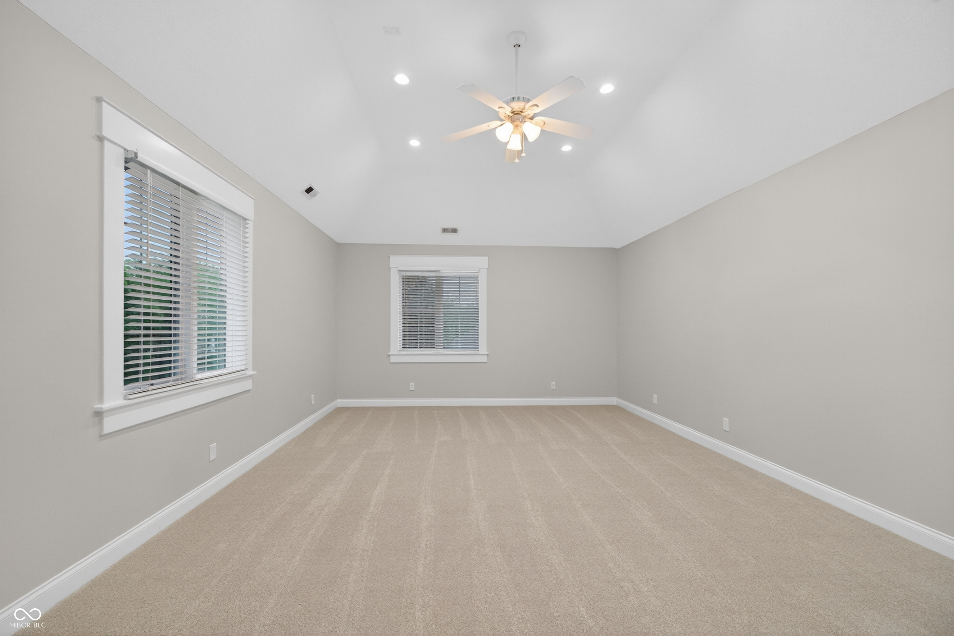 Empty carpeted room with two windows and a ceiling fan with lights.