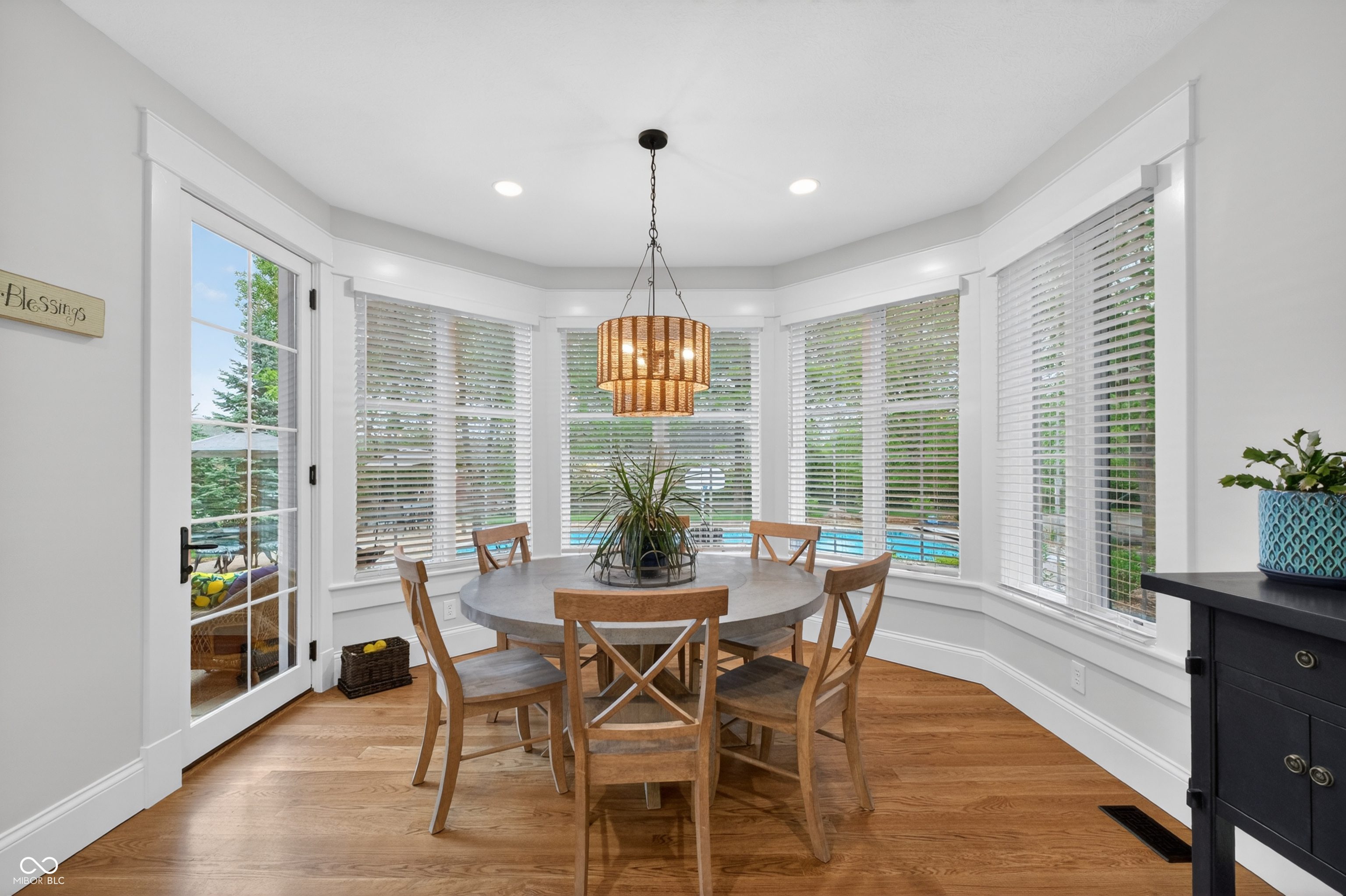 Dining area with a round table and wooden chairs in front of bay windows.