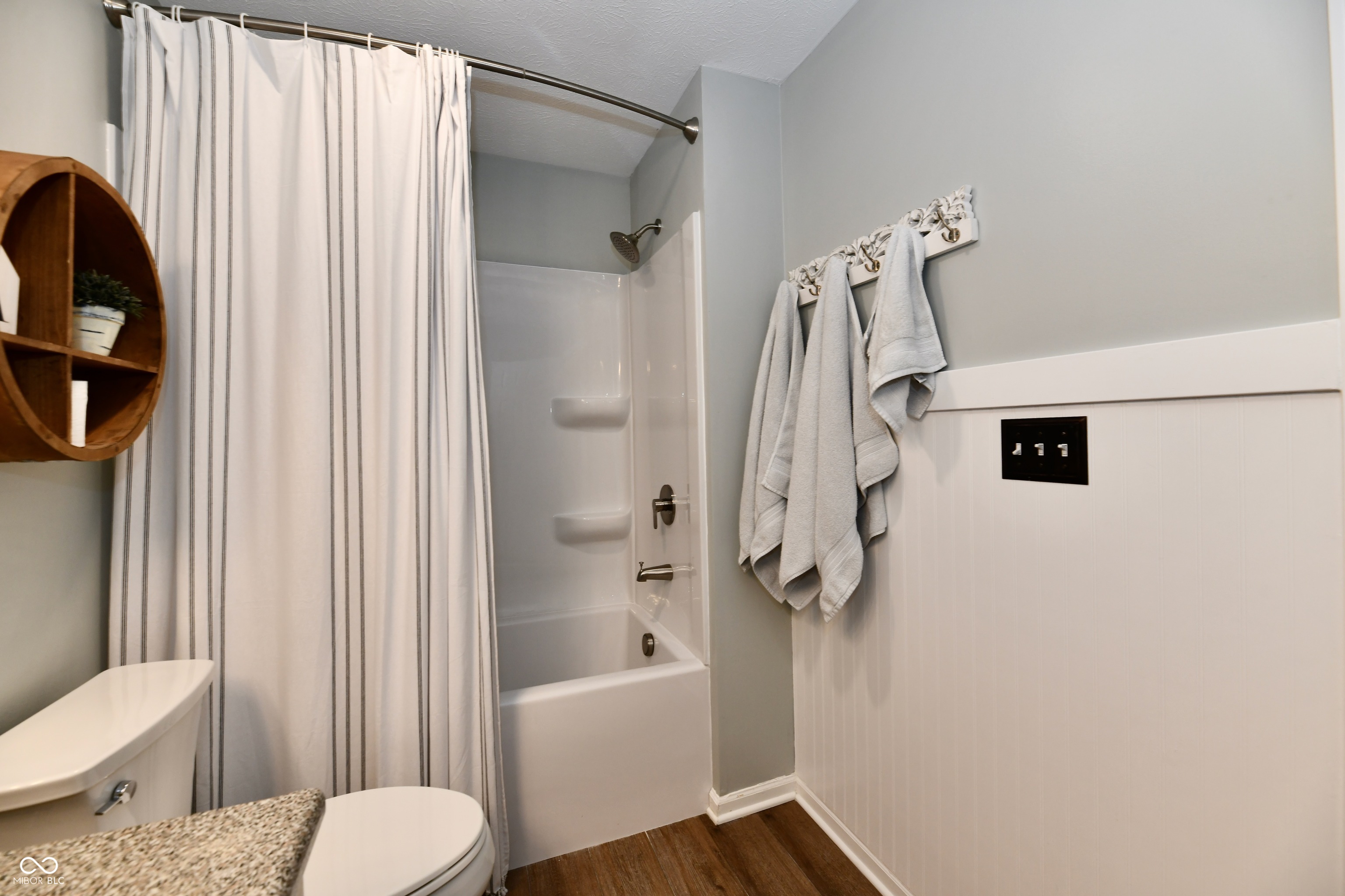 Bathroom with a tub and shower, striped curtain, wall-mounted towels, and wood-look flooring.
