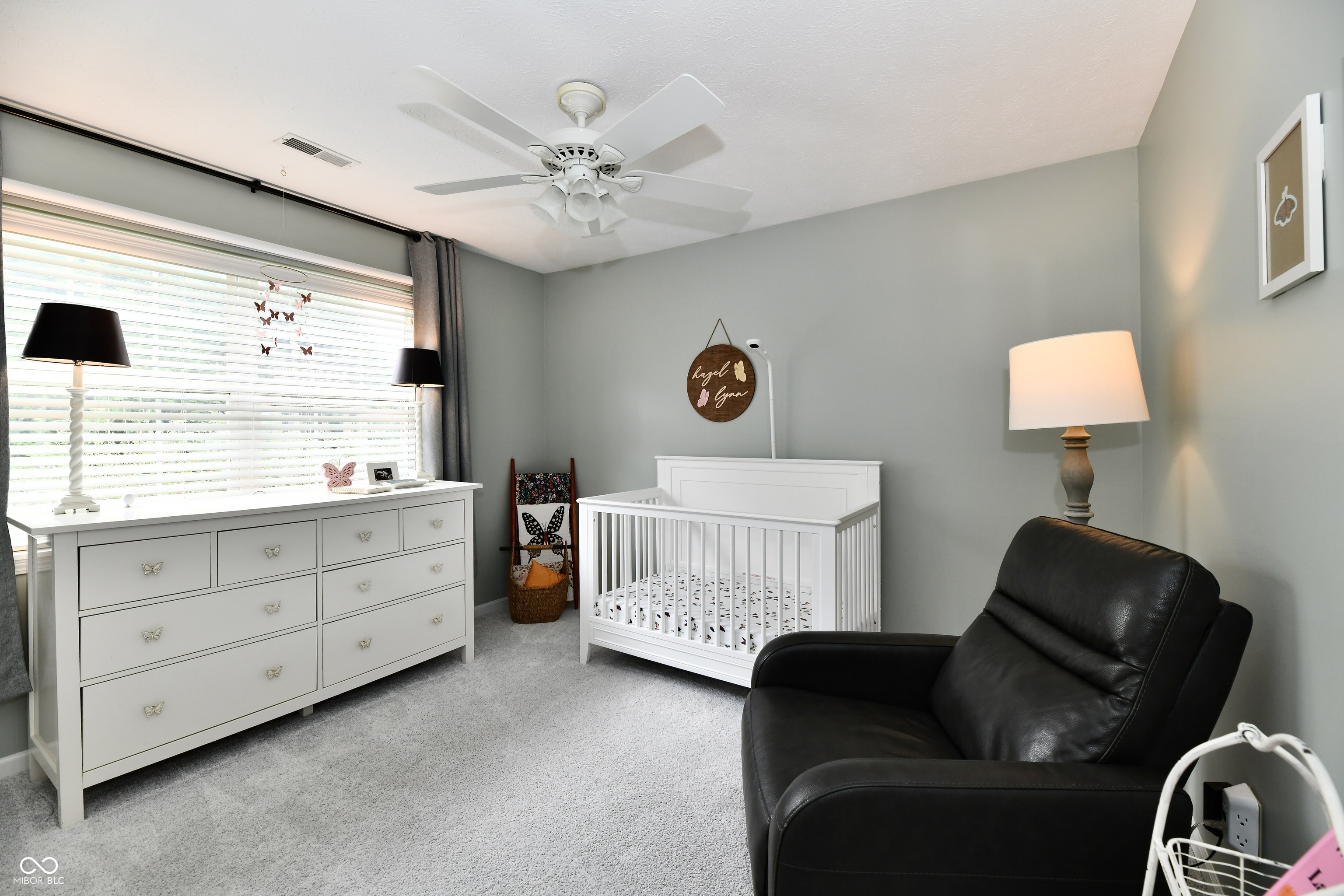 Nursery with a white crib and dresser, a large window, and a black leather glider.